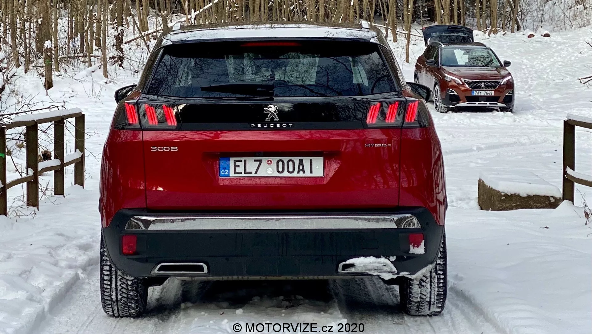 A kép egy vörös Peugeot 3008 SUV hátulnézetét mutatja alsó szögből, kiemelve jellegzetes háromcsíkos karom hátsó lámpáit és központi Peugeot Oroszlán emblémáját, valamint két tónusú lökhárítóját. A jármű havas talajon áll, ami téli körülményeket sugall. A látható jellemzők közé tartoznak az érzékelők a hátsó lökhárítóban, a hátsó ablaktörlő és a hátsó antenna, amely a vezetőtámogató technológiát, valamint a GPS/rádió potenciális funkcionalitását mutatja, ideális autótervezési, biztonsági funkciókkal kapcsolatos és téli vezetési beszélgetésekhez.