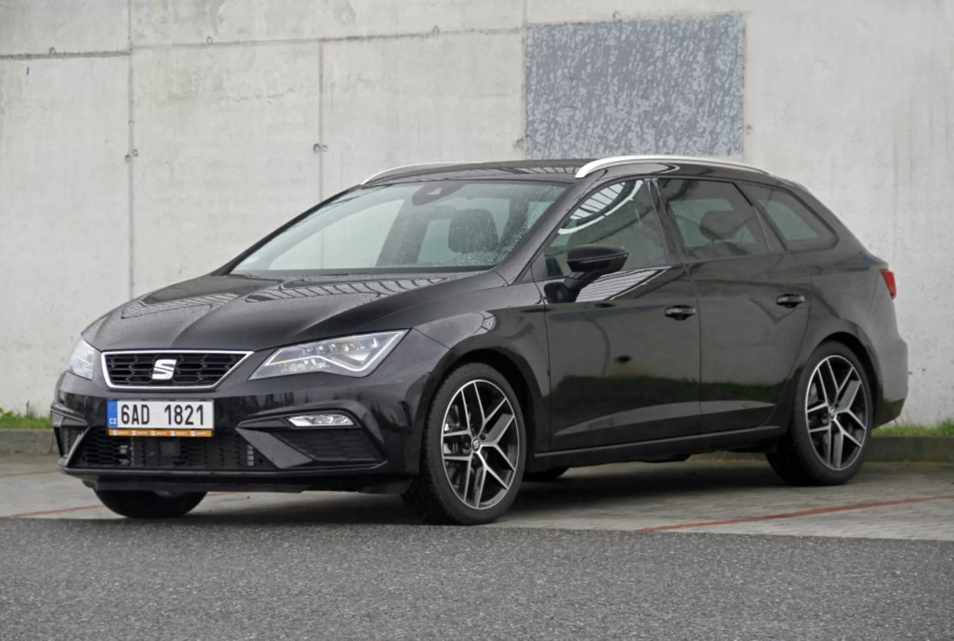 2013-as sötétszínű Peugeot 308 SW hatchback jármű, amely bemutatja jellemzőit: keskeny hűtőrács emblémával, elegáns fényszóróegységek nappali menetfényekkel, sportos többsugaras ötvözött kerekek, jellegzetes hatchback oldalprofil lejtő tetővonallal, kontrasztos sötét karosszériaszínben, háromnegyedes első szögből fotózva ipari háttérrel