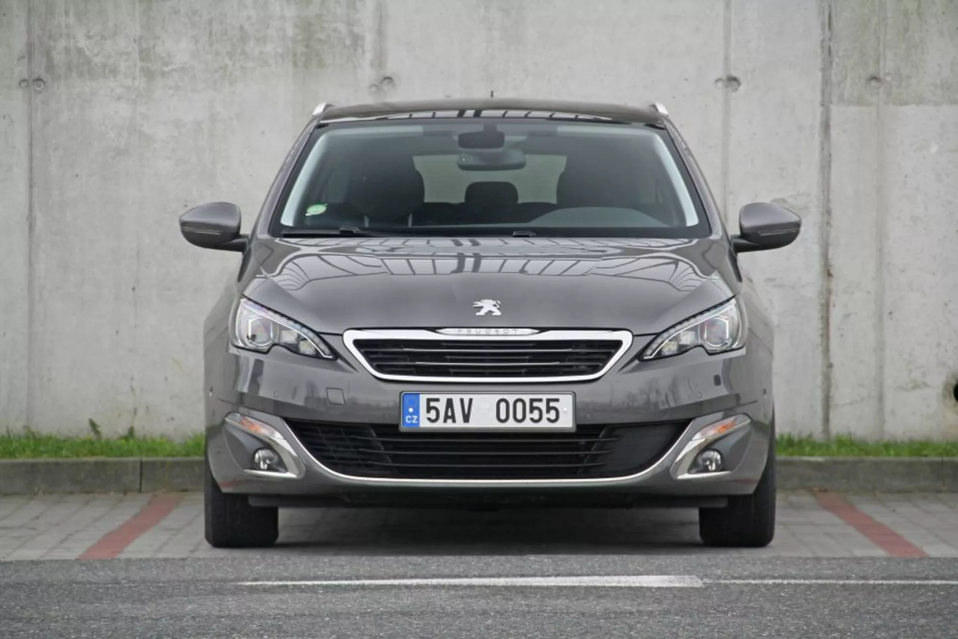 Egy 2013-as Peugeot 308 SW eleje sötétszürke színben, krómozott ráccsal, elegáns fényszórókkal, ködlámpákkal, sportos első lökhárítóval, enyhe kapucnis redőkkel, látható ablaktörlőkkel, európai rendszámtáblával, parkoló szenzorokkal, méhsejtes alsó ráccsal és látható panoráma tetővel, parkolóbeton területen akadályok nélkül