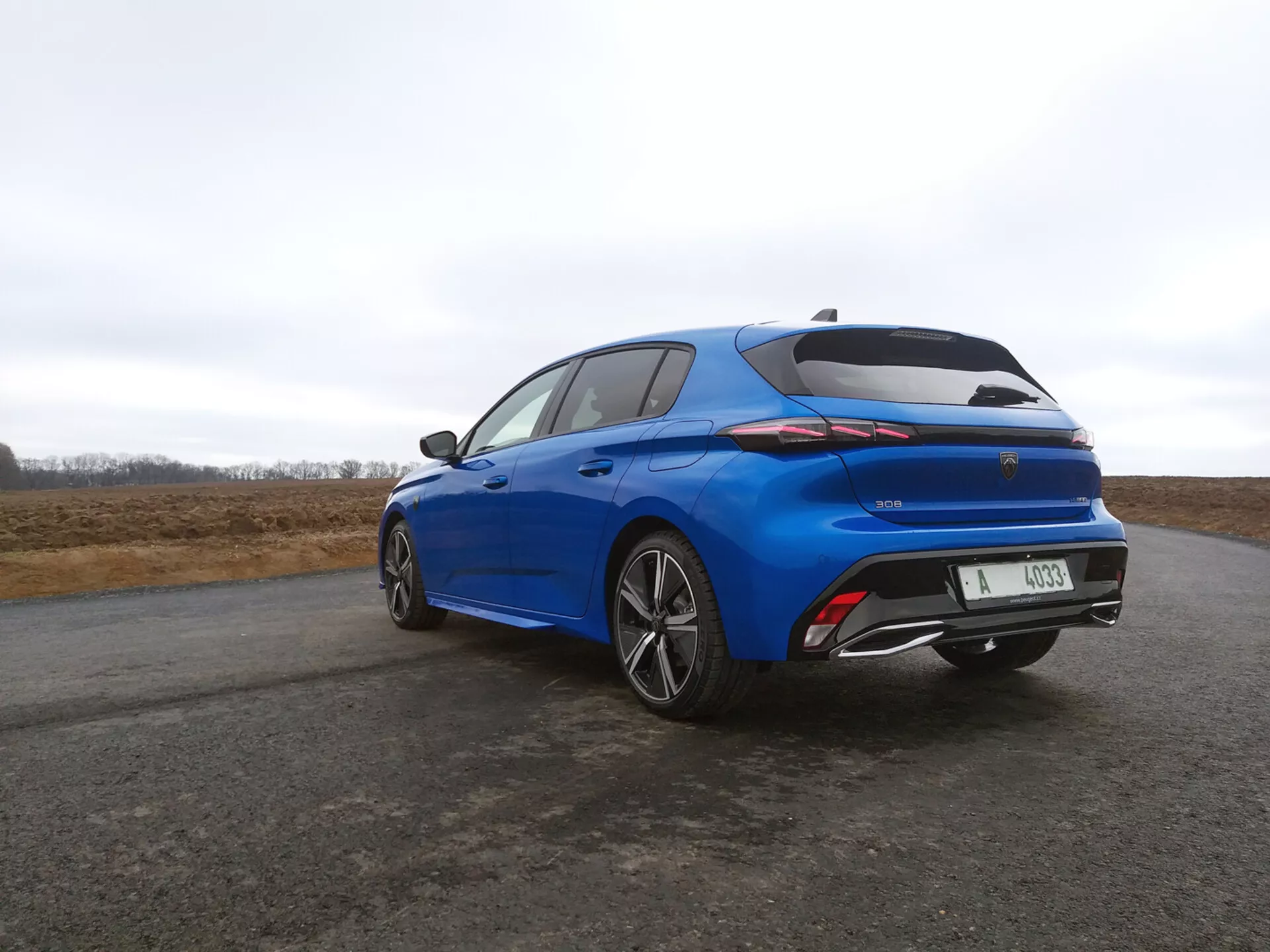 2022-es Peugeot 308 hatchback élénk kékben, hátsó háromnegyedes nézettel, amely bemutatja a LED hátsó lámpákat, integrált spoiler-t, és sok küllős ötvözet kerekeket borús égbolt ellen.