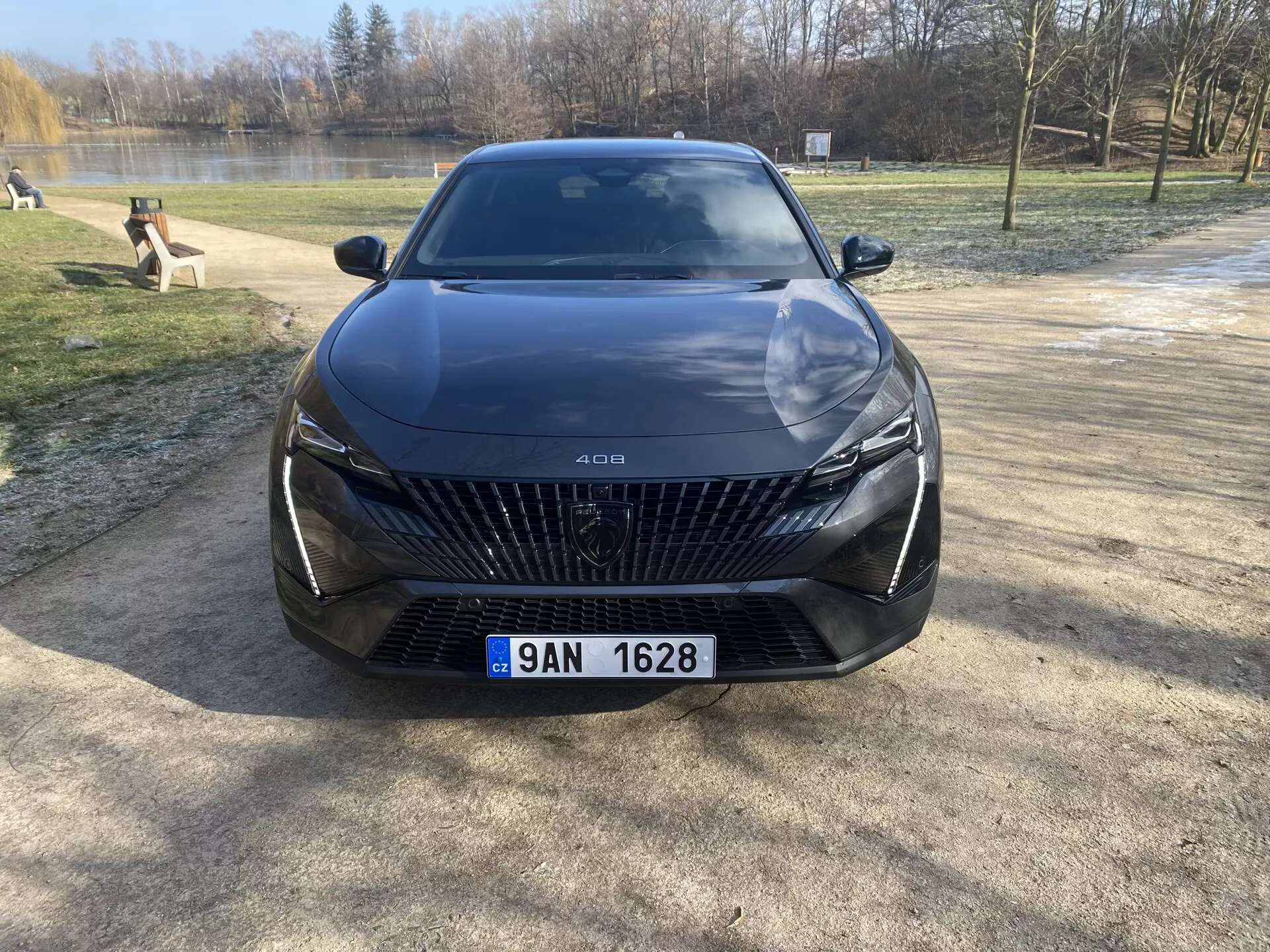 2023-as Peugeot 408 első nézete, kavicsos talajon parkolva, fák és vízterület háttérrel, kiemelve a nagy rácsot gyémánt alakú mintákkal, elegáns LED fényszórókat, szobrászati vonalakat a motorháztetőn és fejlett vezetői asszisztencia funkciókat