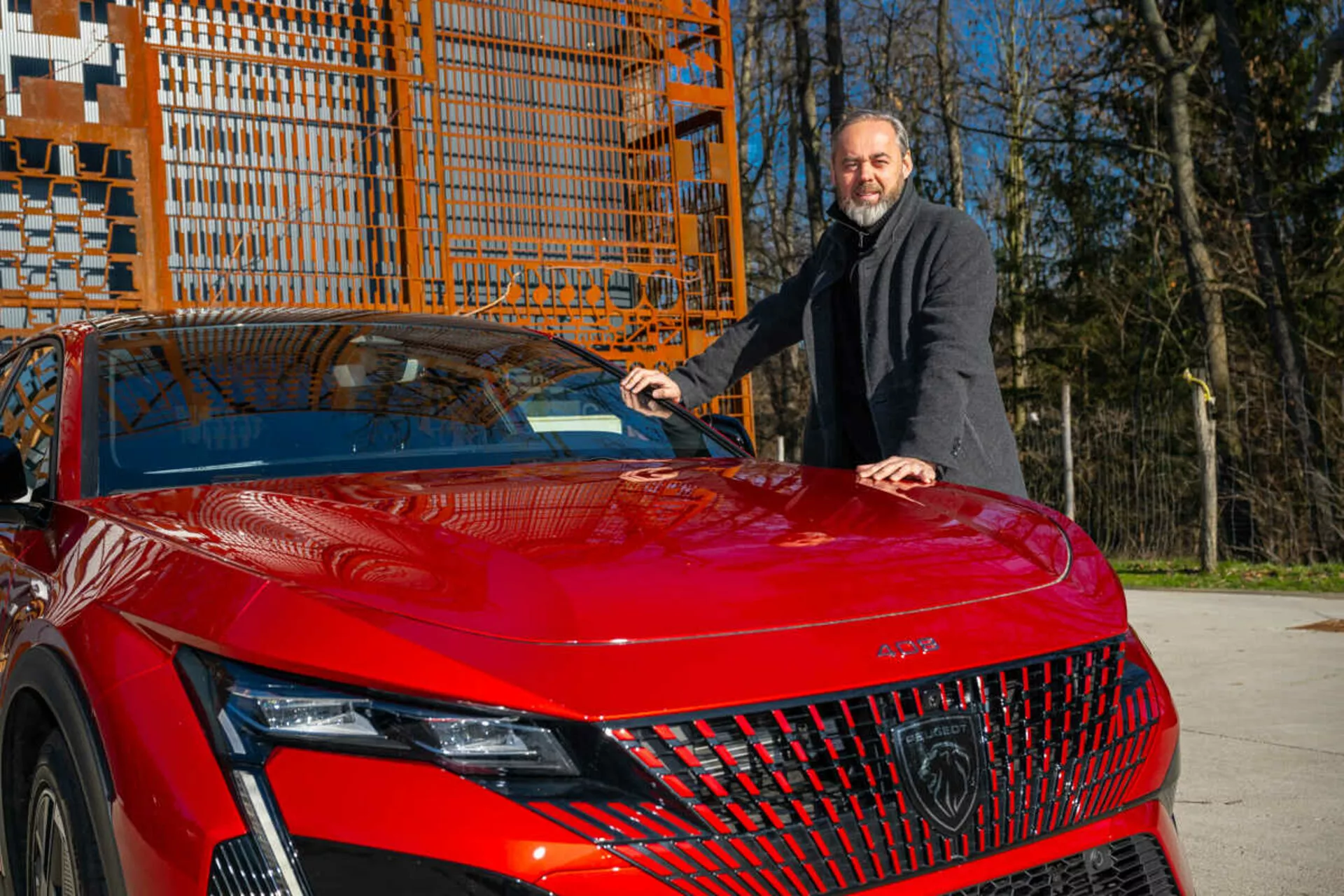 2023-as Piros Peugeot 408 modell bemutatása a napfényben, az elől bal oldali negyedét érzékeltetve, kiemelve a részletes LED fényszórókat, geometrikus hűtőrácsot és sportos, aerodinamikus motorháztetőt, mellette álló, sötét kabátban lévő férfival, aki a kezét a motorháztetőre helyezi