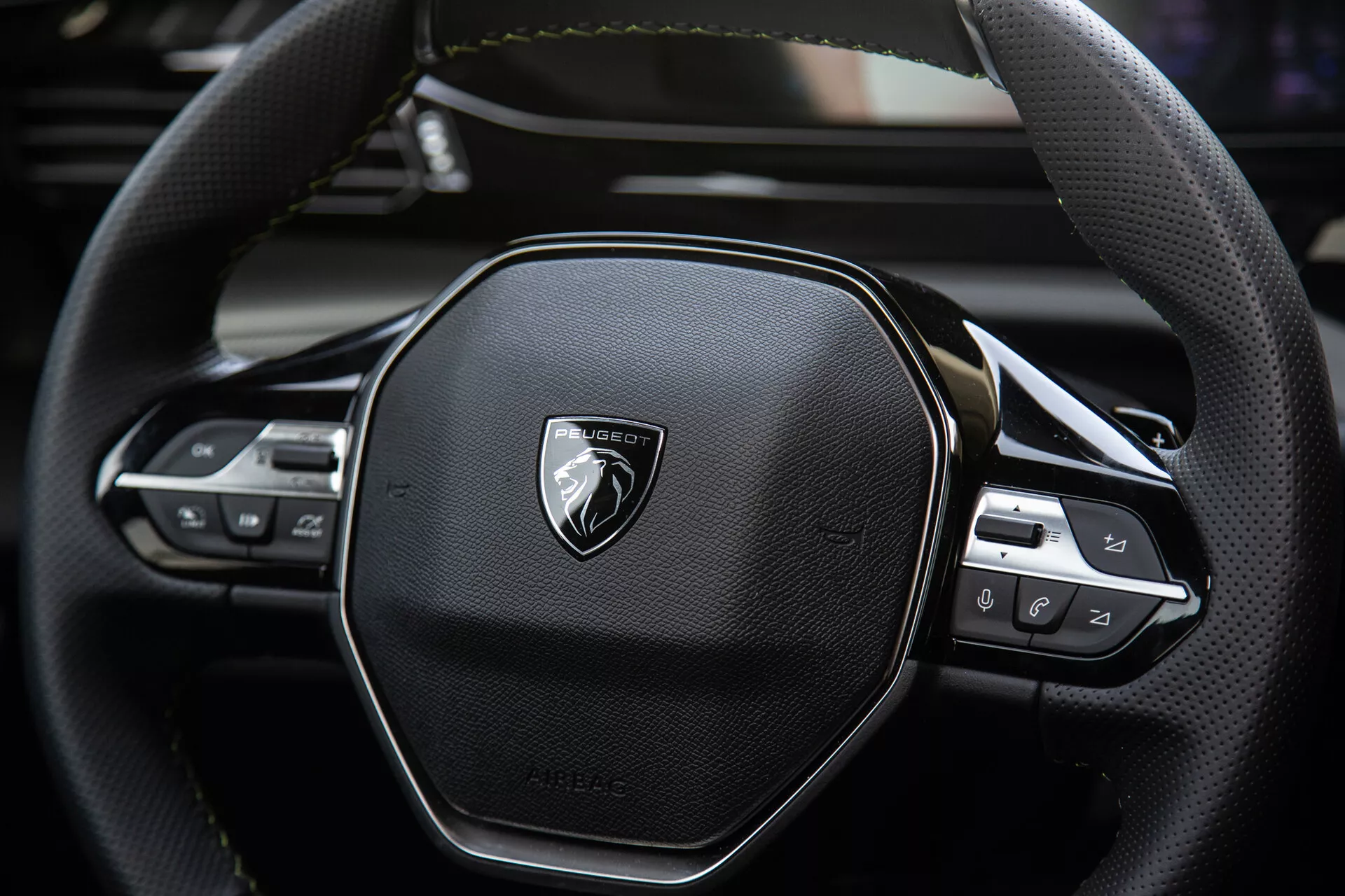 Közeli nézet a Peugeot 408 2023 kormánykerékre, amely bemutatja az ergonomikus hatszögletű kialakítást, a bőrvarrást, az audio és hajózási beállítások integrált technológiai vezérlőit, emblémával a kefélt fémeken.