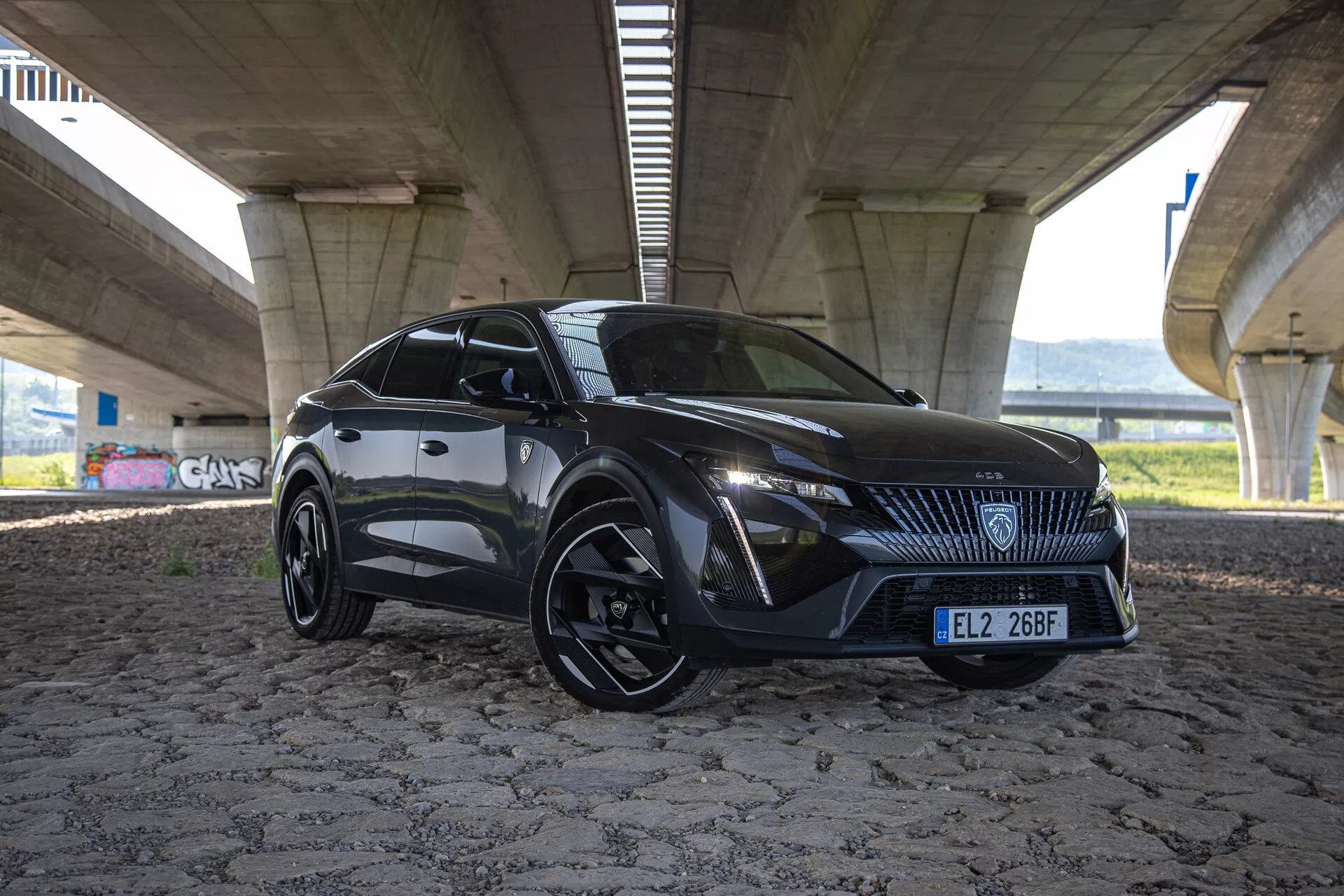 2023-as Peugeot 408 dinamikus pózban egy híd alatt, bemutatva sportos tervezését szögletes vonalakkal, kiemelkedő első hűtőráccsal központi jelvénnyel, éles LED fényszórókkal, aerodinamikus kontúrokkal a motorháztetőn és alacsony profilú, többsugáras kerekekkel, betekintést nyújtva a modern autótervezésbe és technológiába