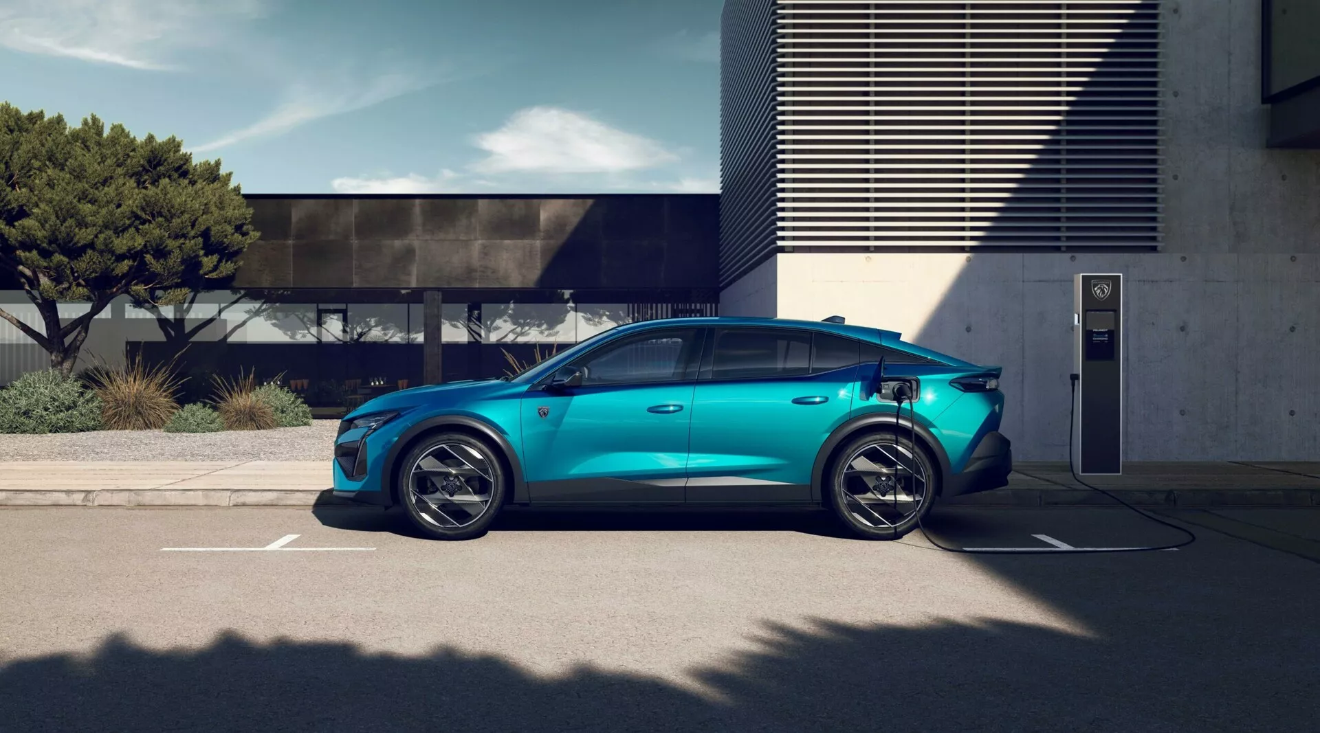 2023-as Peugeot 408 elektromos jármű metálkék színben, kültéri töltőállomáson parkolva, bemutatva sportos ötkarú ötvözött kerekeket, aerodinamikai sziluettet, süllyesztett ajtókilincseket, látható töltőportot, hagyományos oldalsó tükröket, privát üvegezést és panoráma napfénytetőt világos nappali fényviszonyok mellett modern építészettel a háttérben