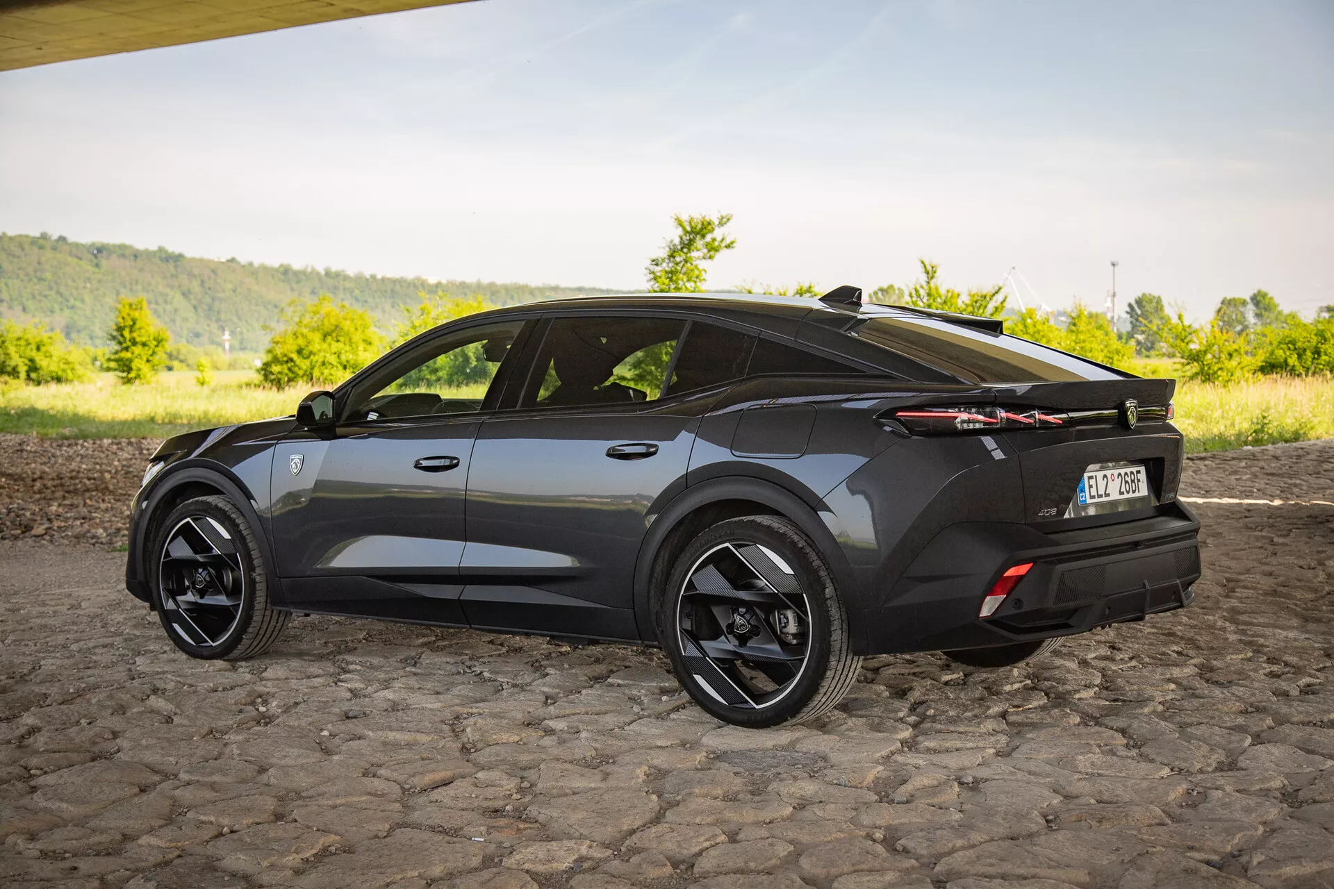 2023-as Peugeot 408 crossover SUV sötét kivitelben, háromnegyedes hátsó nézetben ábrázolva, kiemelve az aerodinamikai kialakítást éles szögekkel, sportos lejtős tetővonallal, nagy fekete ötvözött kerekekkel, jellegzetes LED hátsó lámpákkal és egy finom hátsó spoilerrel macskaköves felületen