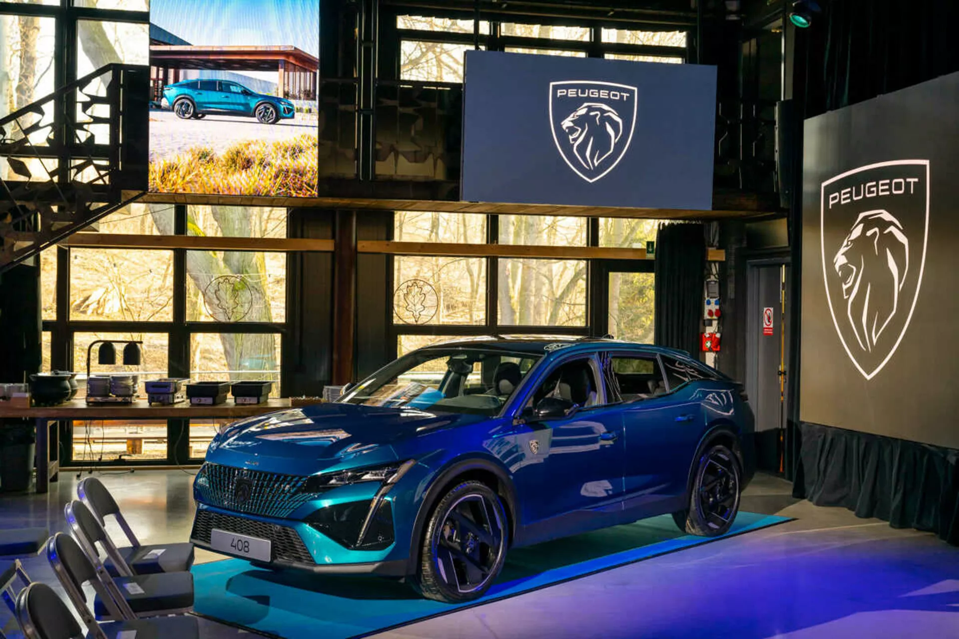 2023-as kék Peugeot 408, belső eseményen bemutatva, jellegzetes hűtőráccsal, éles LED fényszórókkal, sportosan kontúrozott motorháztetővel és légbeömlőkkel, nagy ötvözetű kerekekkel, és dinamikus oldalprofilnal az ajtón szerelt oldalsó tükrökbe integrált irányjelzőkkel