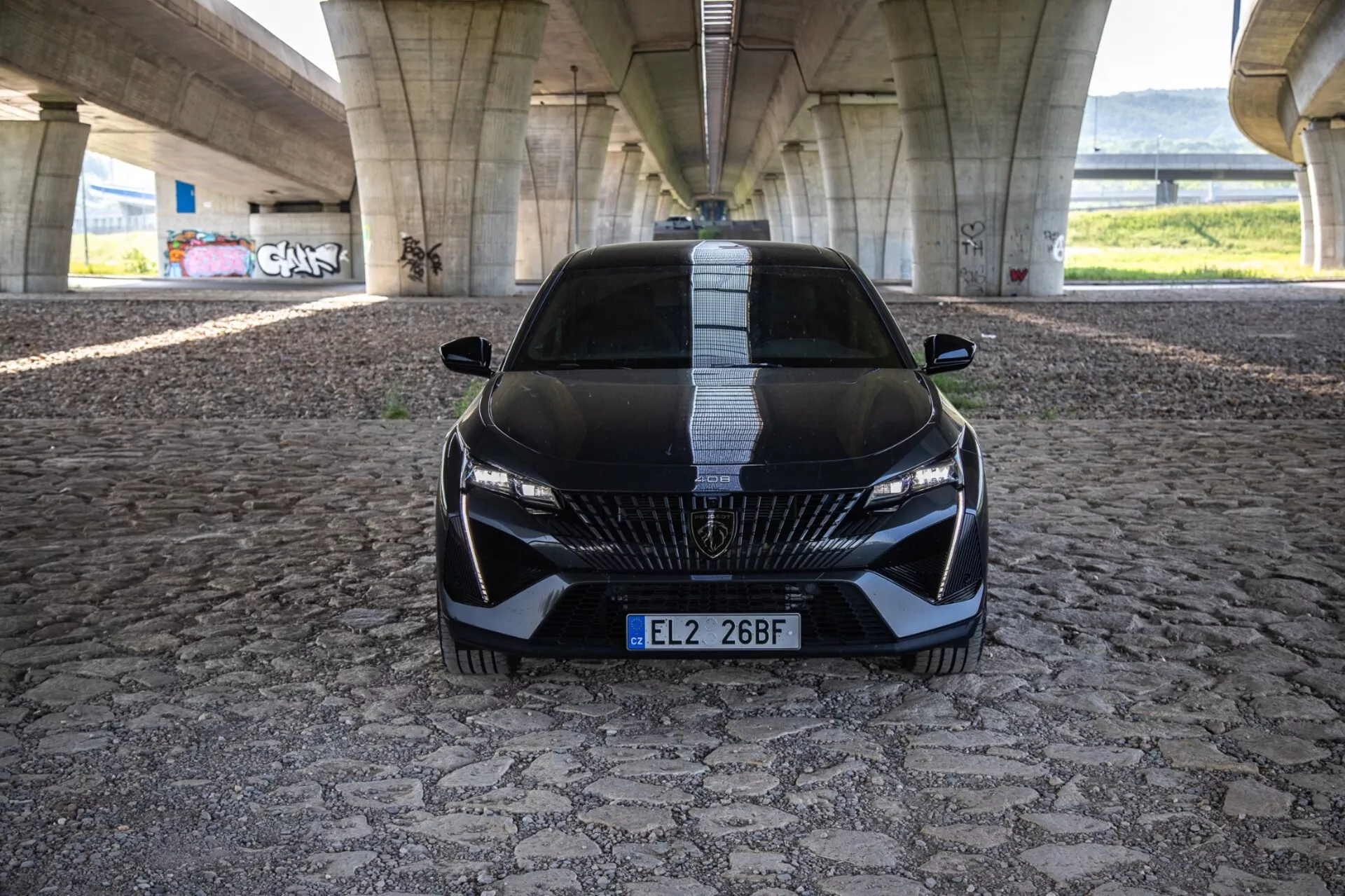 2023-as Peugeot 408 dinamikus front nézetben, amely kiemeli az agresszív tartást és sportos megjelenést részletes rács, elegáns LED fényszórók és aerodinamikai jellemzőkkel mintázott járdán városi híd alatt