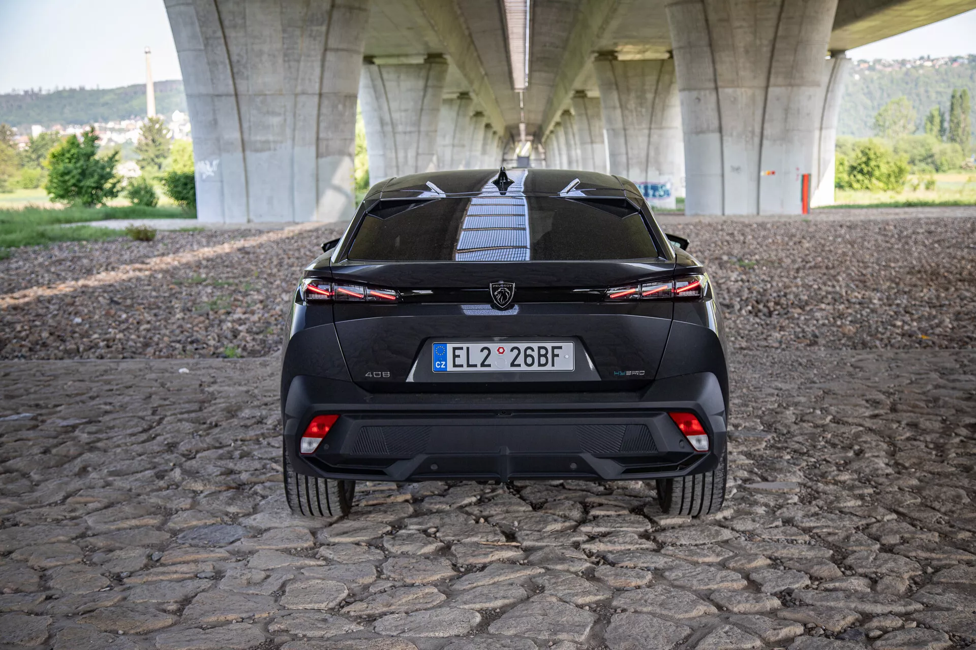 2023-as Peugeot 408 hátsó nézete, amely bemutatja a csomagtartót, lökhárítót, az alsó diffúzort, a szélességet átívelő hátsó lámpákat kapcsoló fénybárral, és parkolást segítő szenzorokat, híd alatt kockaköves felületen fotózva