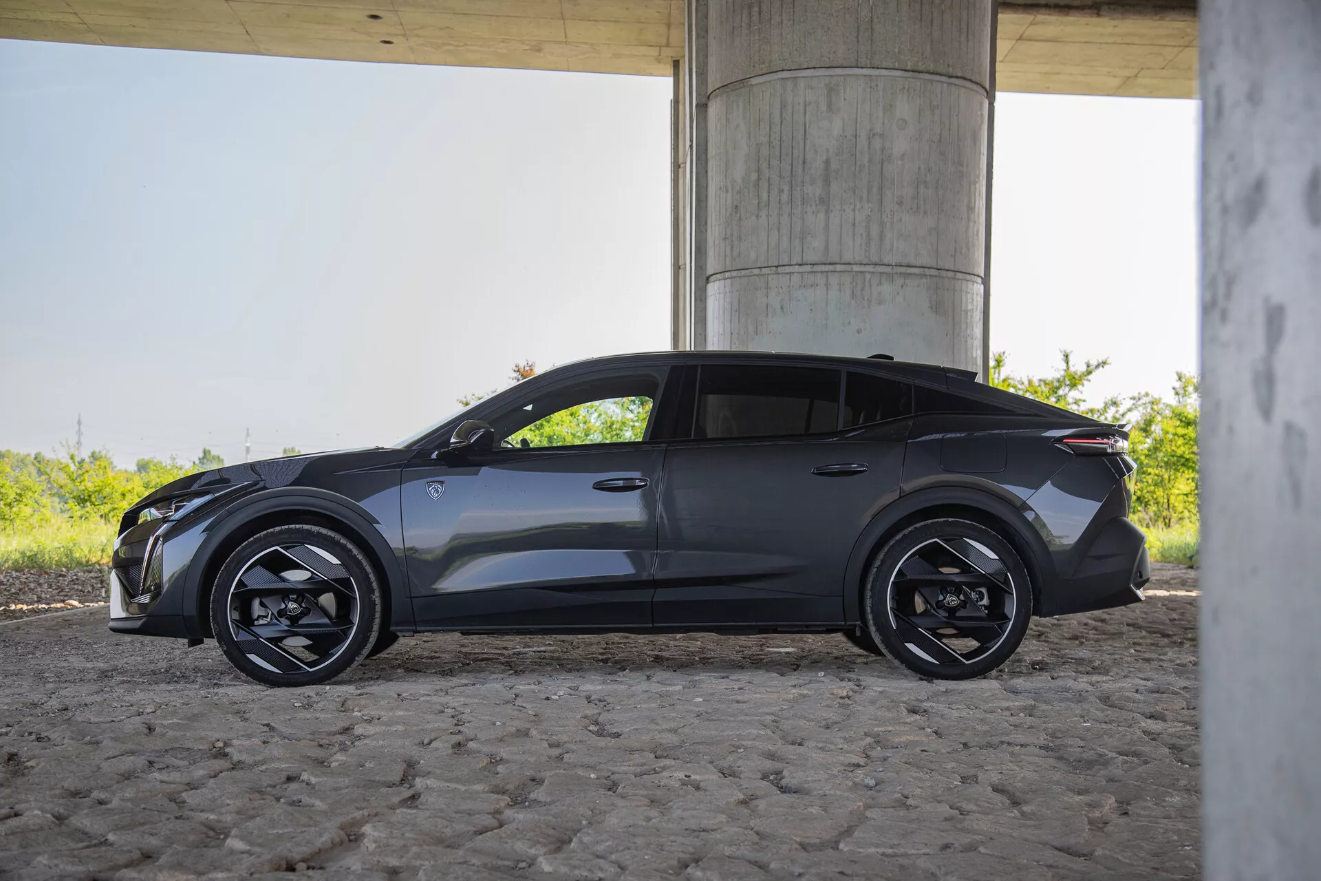2023-as Peugeot 408 oldalnézete kültéren, híd alatt, elegáns tervezéssel, elektromos/hibrid járművekre jellemző zárt hűtőráccsal, szögletes LED fényszórókkal, fekete többspókás kerekekkel, alacsony profilú abroncsokkal, aerodinamikai oldalsó tükrökkel beépített irányjelzőkkel, test színű ajtókilincsekkel, dinamikus karaktervonalal, sportos hátsó rész kompakt spoilerrel, fémes fényezés természetes nappali fényben