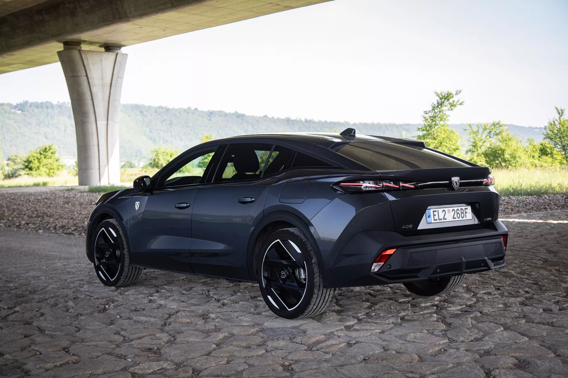 Háromnegyedes hátsó nézet egy 2023-as Peugeot 408 hatchback alatt híd alatt macskaköves úton sportos felnikkel és fejlett világítástechnológiával