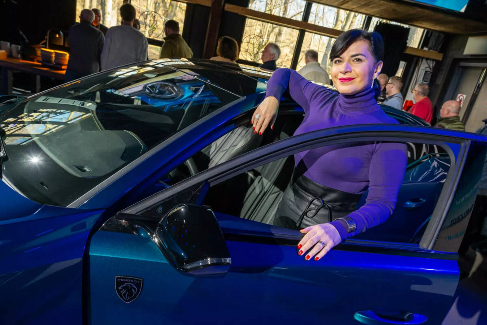 Élénk kék sportautó, valószínűleg egy Lamborghini, háromnegyed felülnézetből fotózva, kiemelve a karosszéria elegáns vonalait, éles LED fényszóróit és jellegzetes pajzs alakú logóját, egy személy lazán érintkezik a járművel, luxust és stílust sugallva.