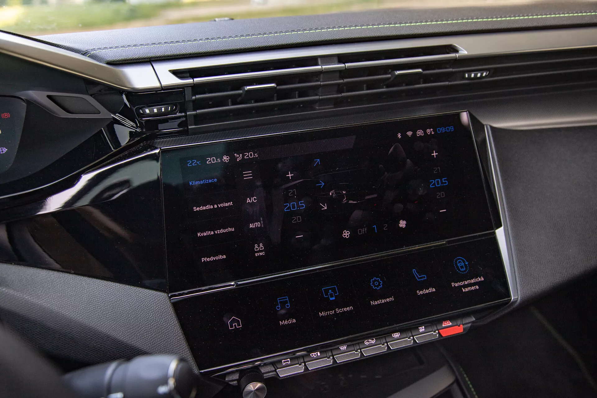 Peugeot 408 2023-as érintőképernyős infotainment rendszerének közelképe, mely 8-10 colos képernyőt mutat navigációval, klímakontrollal, médialejátszás ikonokkal és a műszerfal egy részével, az utas oldaláról készült