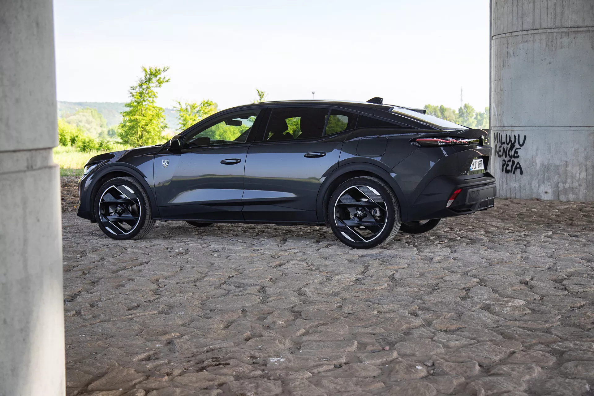 2023-as Peugeot 408 modern, sötétszínű autó oldalnézete híd alatt graffiti-val, aerodinamikus tervezéssel, ötvözött kerekekkel és sötétített ablakokkal