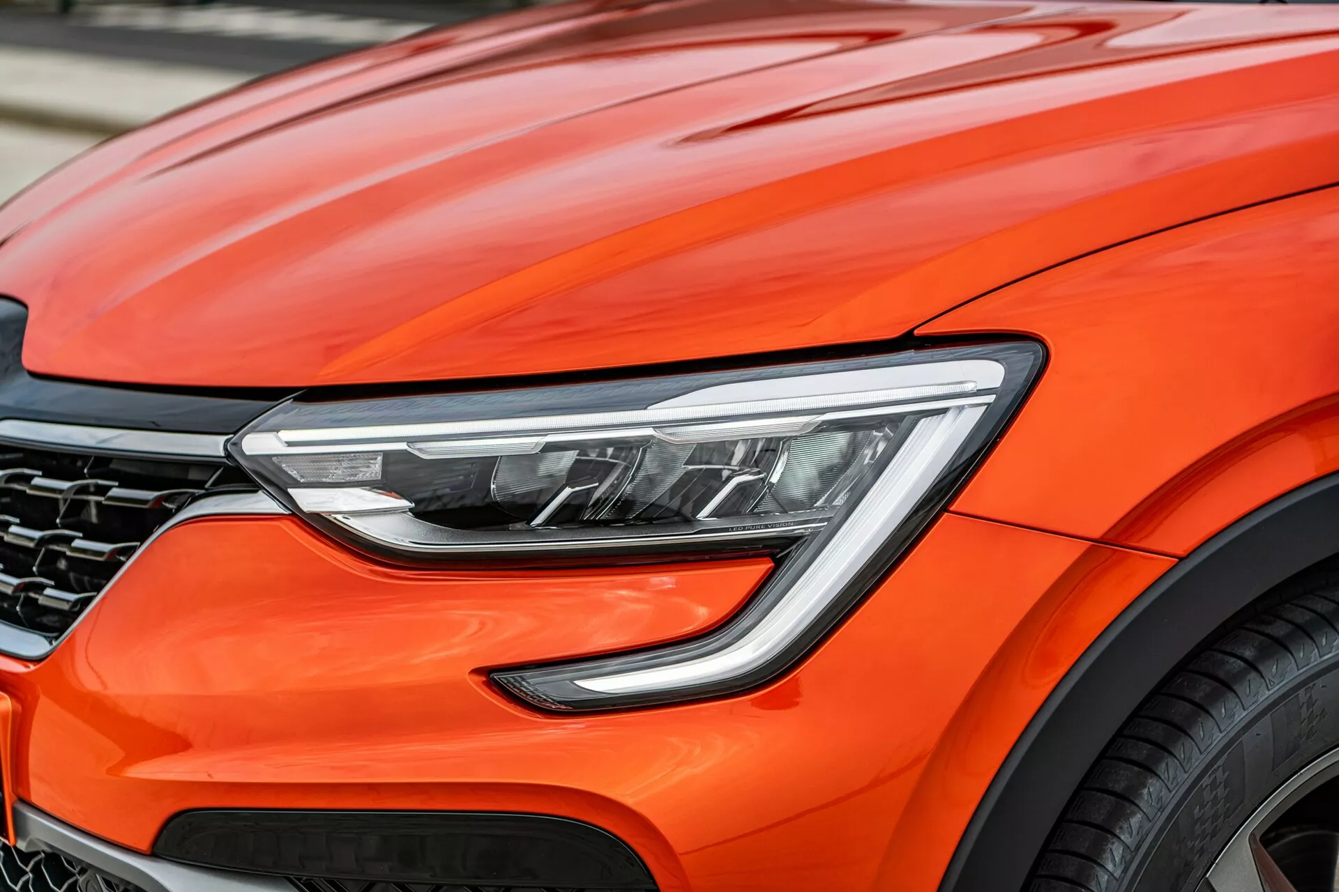 Közelkép egy fényes narancssárga 2021-es Renault Arkana elülső részéről, amely a modern dizájnelemekre összpontosít, beleértve a fejlett LED fényszórókat, a motorháztető aerodinamikus íveit, a részben látható sötét méhsejt rácsot és egy nagy könnyűfém kerék darabját, amely prémium teljesítményt és élvonalbeli autóipari technológiát sugall.