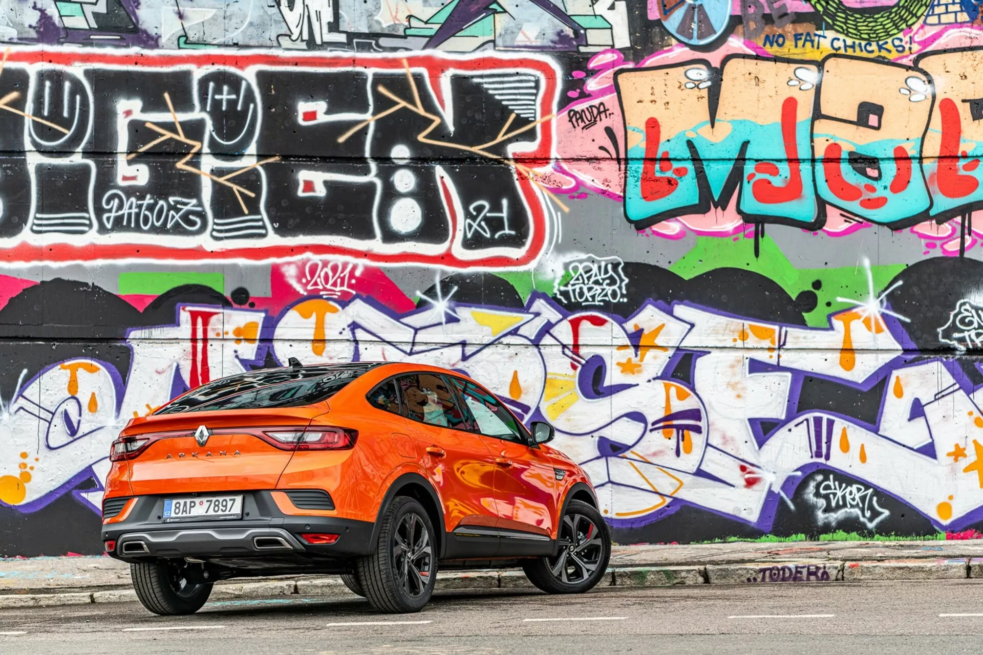 Oldalsó profilnézet egy élénk narancssárga Renault Arkana 2021 crossover SUV-ról, amely városi úton parkol. A jobb oldalról fotografálva és a hátsó háromnegyedre fókuszálva, a kép az autó sportos, lejtős tetővonalát, hátsó légterelőjét, magas övvonalát, nagy, stílusos könnyűfém keréktárcsáit, sötétített ablakait és aerodinamikus jellemvonalait rögzíti. A hátsó lámpák elegánsak és valószínűleg LED-esek. A háttérben egy élénk, graffiti-vel festett fal látható, ami fokozza a városi esztétikát. A nappali fényben készült fotó lágy árnyékokat mutat az autó alatt, kiemelve a dizájn részleteit.