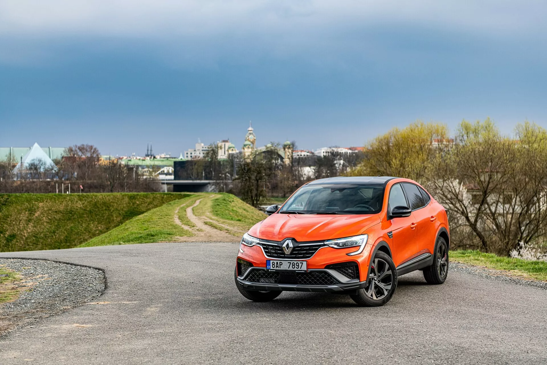 A képen egy modern 2021-es Renault Arkana crossover SUV látható, egy kanyargós ösvényen parkolva. Háromnegyed elölnézetből készült fotó, amely világosan megmutatja a jármű elejét a középen elhelyezkedő, hangsúlyos emblémával rendelkező hűtőrácsot, a hátrafelé nyúló, letisztult fényszórókat és a valószínűleg ködlámpákkal és szenzorokkal ellátott, precízen kidolgozott első lökhárítót. Az aerodinamikus tervezésű, esetleges beépített irányjelzőkkel rendelkező oldalsó tükrök, az alufelnik és a vibráló narancsszínű karosszéria teljes megjelenést kölcsönöznek. A háttérben fák és felhős égbolt látható, távolabb pedig épületek, amelyek városi környezetre utalnak.