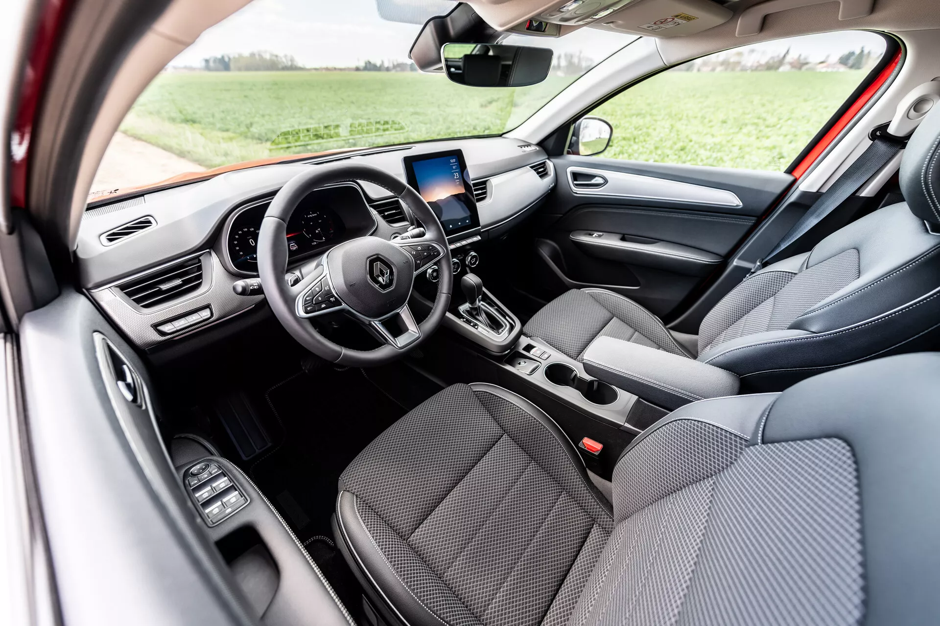 2021-es Renault Arkana belső terének megjelenítése, amely szürke szövetből készült első üléseket, integrált vezérlőgombokkal ellátott multifunkciós háromküllős kormánykereket, digitális műszerfalat, GPS navigációs infotainment képernyőt a központi légbeömlők felett, kézi légkondicionáló vezérlőket, automatikus sebességváltót és az vezető oldali ajtón elhelyezett elektromos ablakvezérlőket mutat, természetes nappali fényben, kültéri környezetben