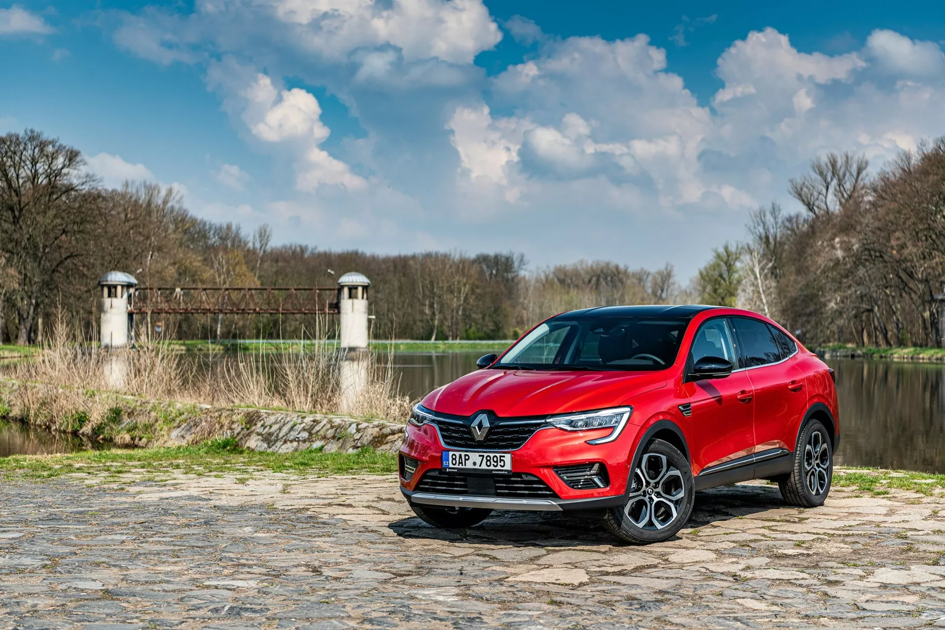 A kép egy 2021-es Renault Arkana-t ábrázol, amely egy macskaköves úton parkol egy folyó vagy tó közelében, növényzettel és háttérben a gát vagy híd rendszerének tornyaihoz hasonló szerkezetekkel. Az autót alacsony szögből, háromnegyed elülső nézetben kapták le, kiemelve a hűtőrácsot, a LED-fényszórókat és az oldalsó profil egy részét, beleértve a vezető ajtaját és az első sárvédőt. A modern dizájnelemek, például az éles LED fényjelzés és a nagy könnyűfém kerekek magasabb felszereltségi szintet javasolnak. A fényvisszaverő fémes festék esztétikai vonzerőt biztosít, míg a vezetőtámogató szenzorok és a lökhárítókba és oldalsó tükrökbe integrált technológia növeli a funkcionalitását.