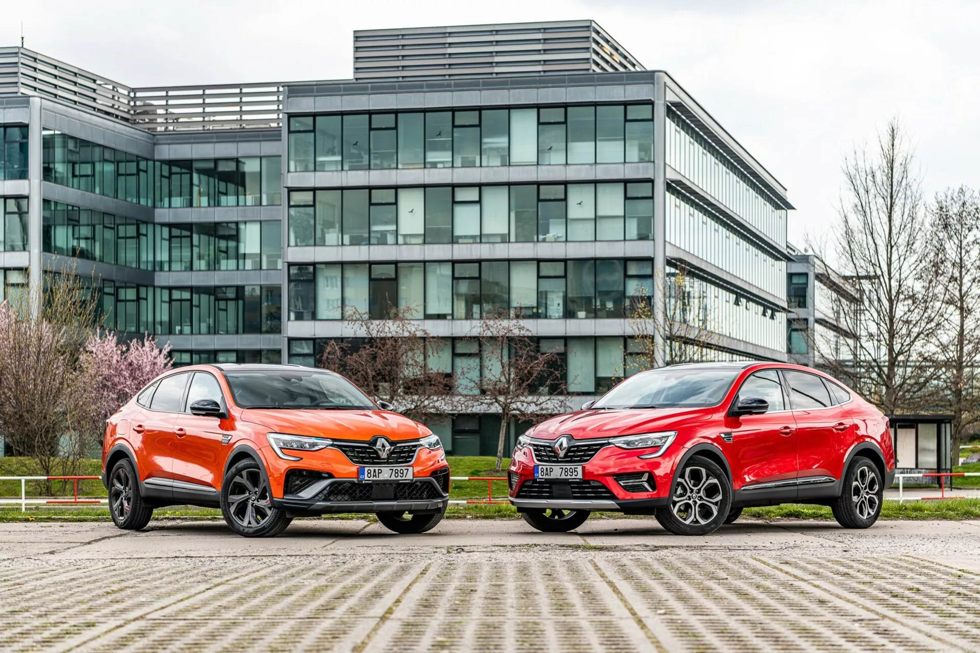 A kép két SUV-stílusú járművet mutat egymás mellett parkolva egy modern üveg homlokzatú irodaház előtt. A bal oldali jármű élénk narancssárga festésű, fémes fényű, és jellegzetes első hűtőrácsa, éles LED fényszórói, faragott motorházfedéllel és nagy, többküllős könnyűfém kerekekkel rendelkezik, alacsony profilú gumiabroncsokkal. A jobb oldali autó fémes piros színű, hasonló összsiluettel, éles fényszórókkal, kiemelkedő hűtőrácsokkal és stilizált nagy könnyűfém kerekekkel. Mindkét járműben szögletes vonalak, aerodinamikus tükrök és kontrasztos alsó karosszériaelemek találhatók. Az alacsony szögből, talajszinthez közel készített kép kiemeli a városi környezetet, és hangsúlyozza a járművek kialakítását, jelenlétét és fejlett technológiai jellemzőit.