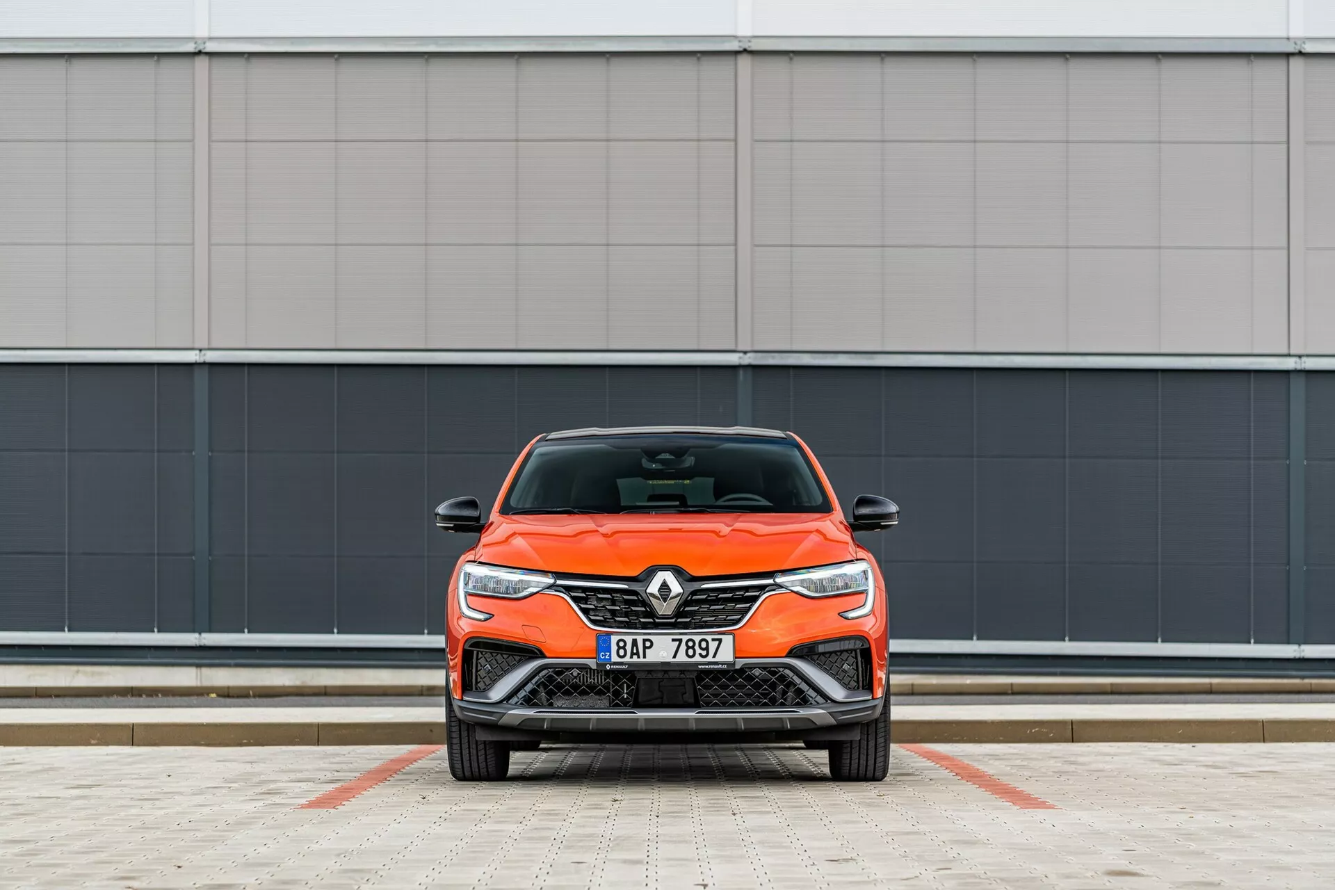 Élénk narancssárga Renault autó frontális nézete, kiemelve annak modern dizájnját elegáns fényszórókkal és első hűtőrácsokkal, merőlegesen parkolva a kamerára egy minimalista szürke fal háttere előtt egy burkolt parkolóhelyen.