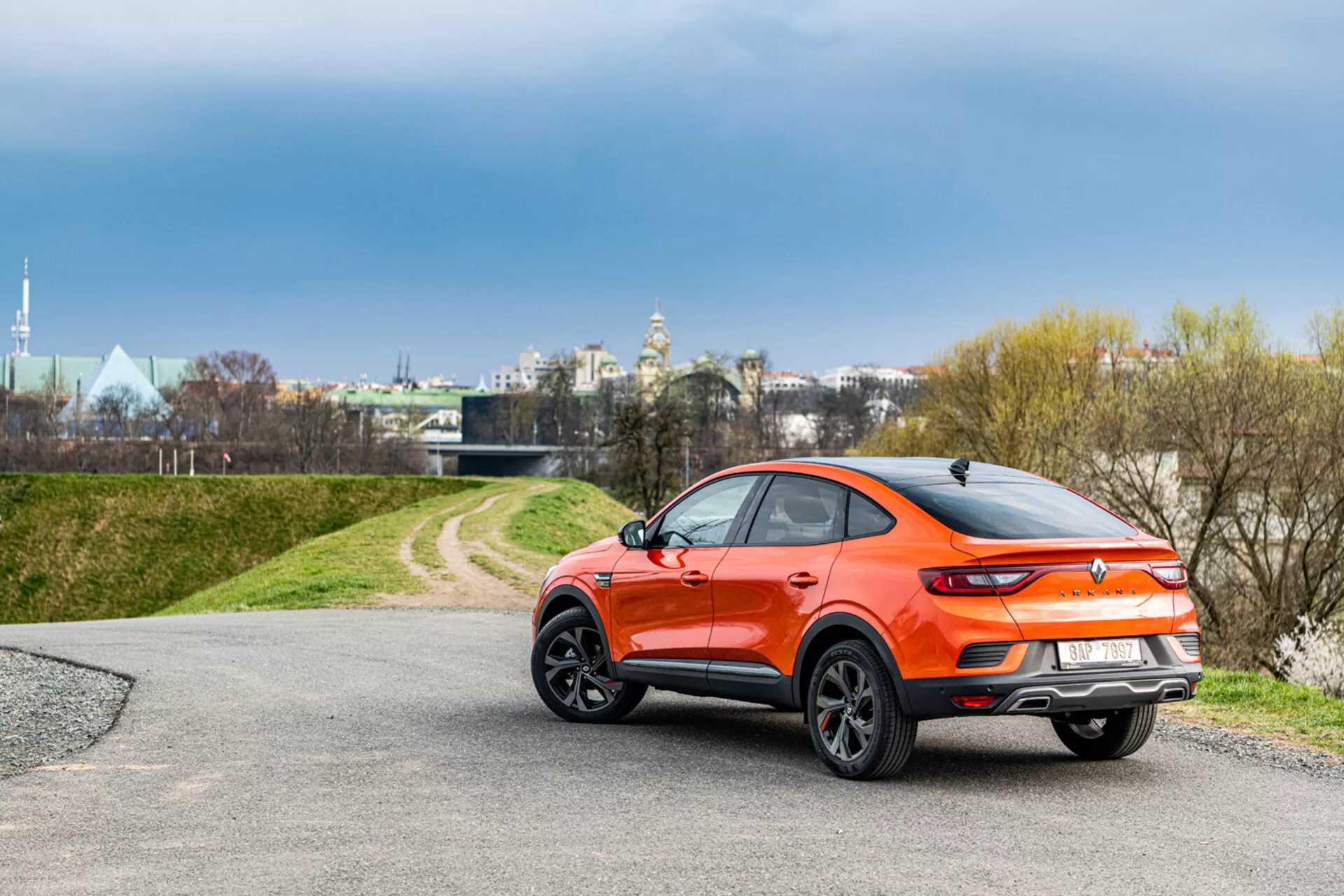 2021-es Renault Arkana narancssárga kompakt crossover SUV, jobb szögben, bemutatva a profilját és a részleges hátsó nézetet, természetes háttér előtt lágy megvilágítással, amely kiemeli sportos kupé-szerű elegáns tervezését és modern jellemzőit.