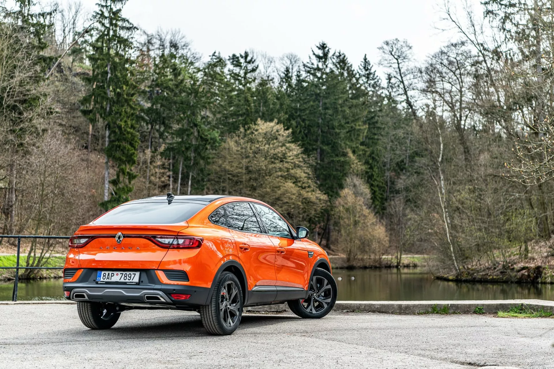 A 2021-es Renault Arkana hátsó háromnegyed szögből ábrázolva, részletes oldal- és hátoldali nézet, modern crossover/SUV, megnövelt hasmagassággal és sportos coupé-szerű tetővonallal. Az élénk narancssárga karosszéria kiemelkedik a fák és a tó hátterében, kiemelve a többrudas designú könnyűfém felniket, a LED hátsó lámpákat, a hátsó spoilert, a cápauszony antennát, a középen elhelyezett emblémát, a kettős kipufogócsövet, a fényes fényezést és a hátsó sötétített üvegeket. A szórt természetes fény javítja a gondozott külső láthatóságát, amit az aktív életmódot kedvelő fogyasztók számára tesz vonzóvá, kikövezett felületen parkolva.