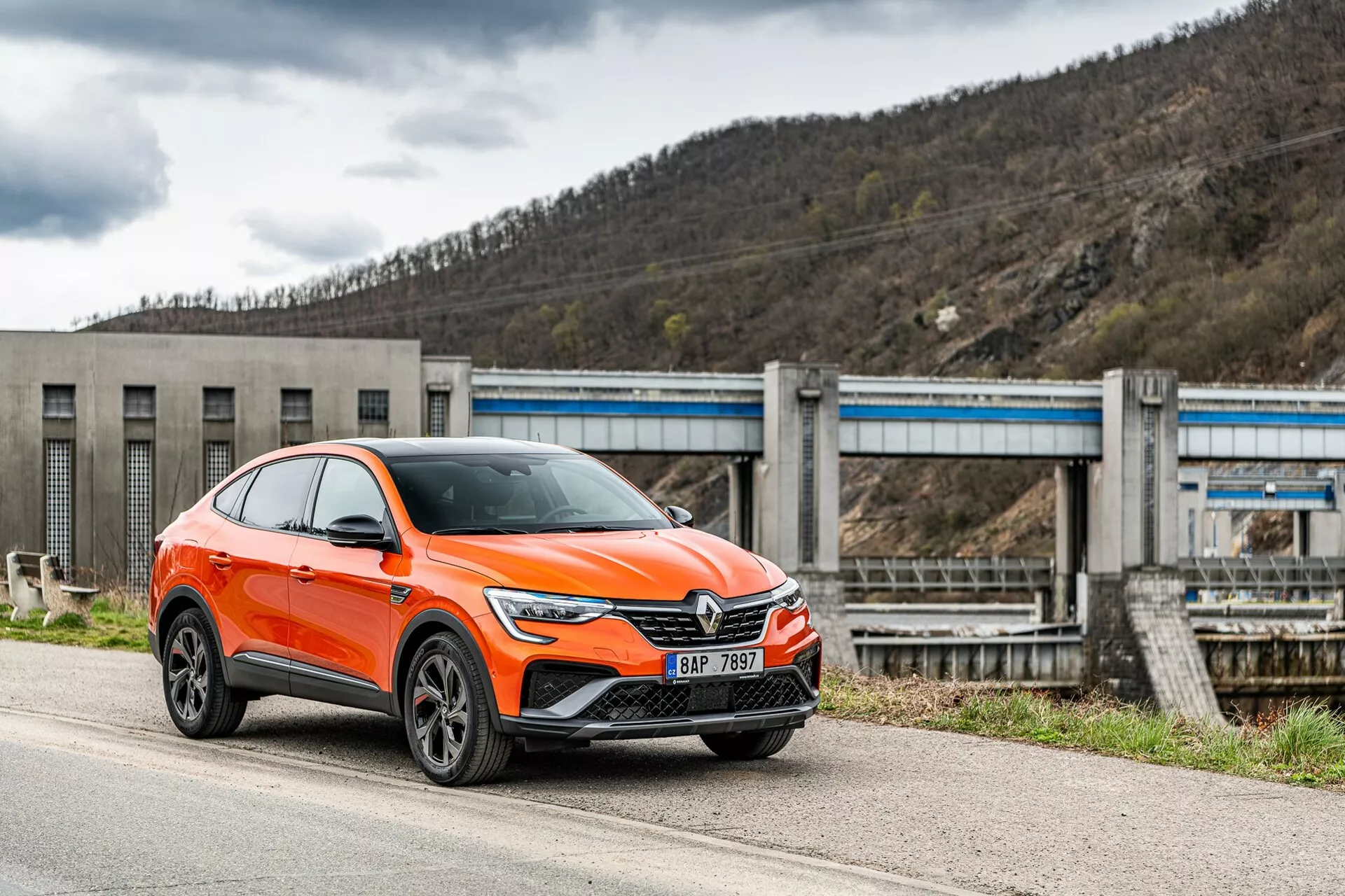 Egy modern, élénk narancssárga Renault Arkana 2021-es crossover SUV parkol az út mentén, elölről oldalról fényképezve, a vezető oldalára összpontosítva. A kép bemutatja az autó első rácsát egy nagy emblémával, elegáns, szögletes LED fényszórókkal, nappali menetfényekkel, légbeömlőkkel és valószínűleg vezetősegítő rendszerek érzékelőivel az alsó lökhárítón, kiemelkedő sárvédőívekkel, többrudas dizájnú könnyűfém kerekekkel és integrált irányjelzőkkel ellátott tükrökkel. Az SUV kategóriába sorolt jármű magasabb állású, nagyobb hasmagassággal és egy kupé stílusú hátrafelé lejtő tetővel rendelkezik. A háttérben egy gát vagy zsiliprendszernek tűnő infrastruktúra látható, amely az autót ipari vagy vidéki környezetbe helyezi. A felvétel kissé alacsony szögből készült, kiemelve az autó magasságát és állását.