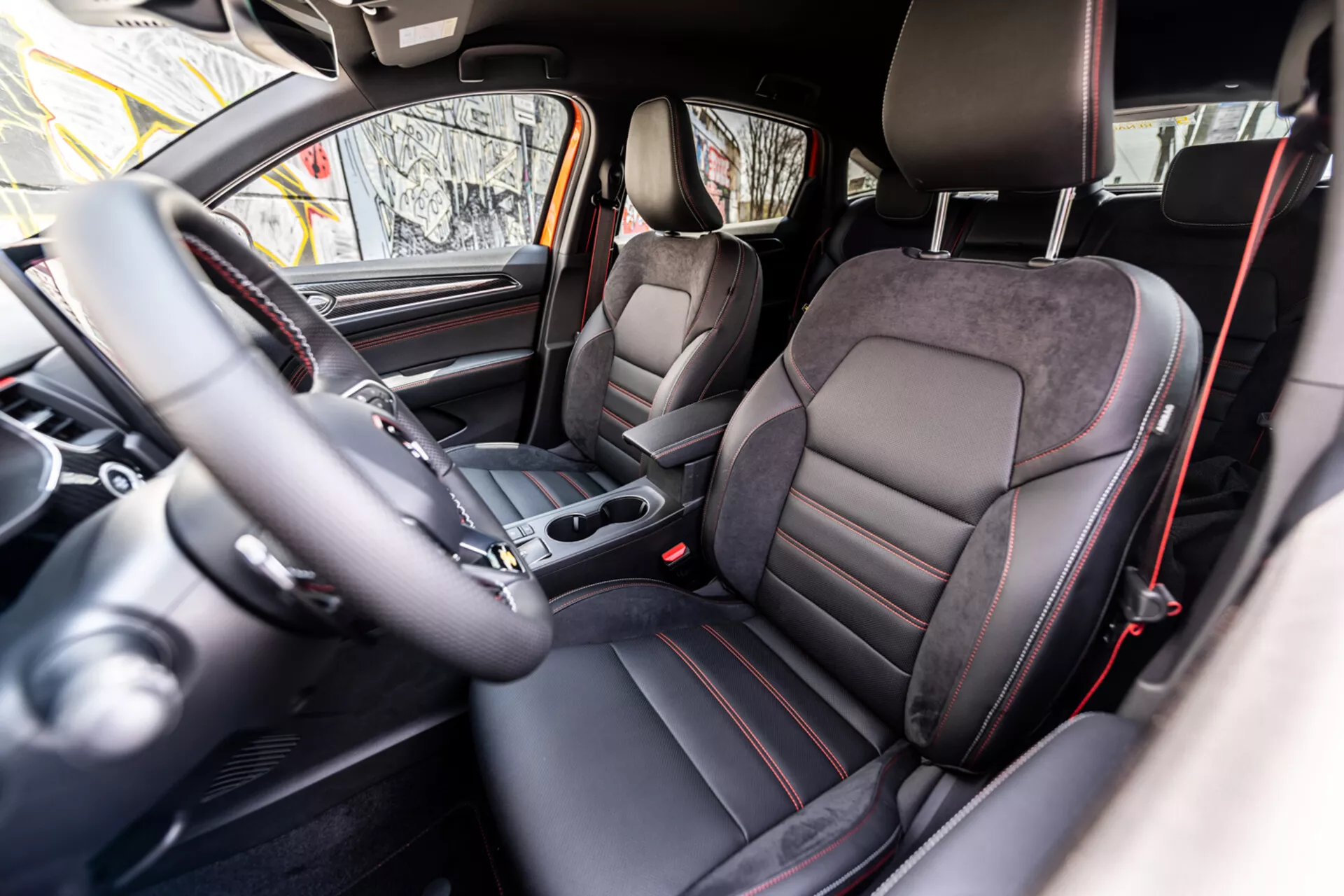 2021-es Renault Arkana belső terének megjelenítése, a hátsó ülésből előre néző perspektívával, sportstílusú, bőr- és Alcantara-borítású ülésekkel, bal oldali kormánnyal szerelt vezérlőkkel, középkonzolon elhelyezett automatikus váltókar, analóg műszerfal, integrált infotainment képernyővel, és prémium sötét színsémával fém díszítőelemekkel