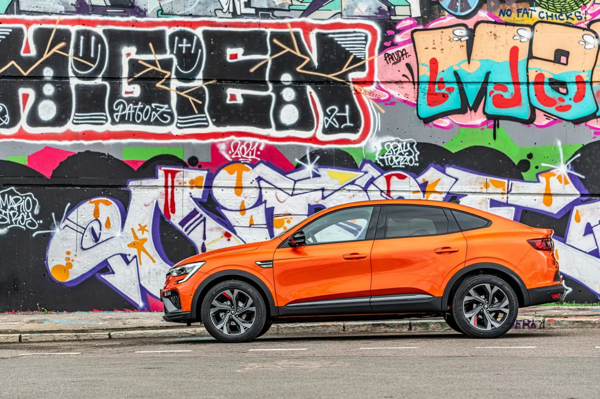 A képen egy élénk narancssárga Renault Arkana 2021 crossover SUV látható, amely párhuzamosan parkol egy utcán, háttérben egy élénk színű graffiti fallal. Az oldalnézetből készült fénykép kiemeli a jármű sportos és modern kialakítását, láthatóak a vezető oldalán lévő első és hátsó ajtók, az oldalsó ablakok, az oldalsó tükrök és a hátrafelé lejtő tetővonal. Az alacsony profilú gumiabroncsokkal ellátott stílusos könnyűfém kerekek az úttestre való teljesítményre utalnak. Az első ajtókon található kulcs nélküli belépő fogantyúk a kulcs nélküli belépésről és indítórendszerről árulkodnak. A kép napfényben készült, kiemelve a narancssárga karosszéria graffiti elleni kontrasztját, és jól karbantartott, éles karosszériát mutat egy fiatalos, városi környezetben.