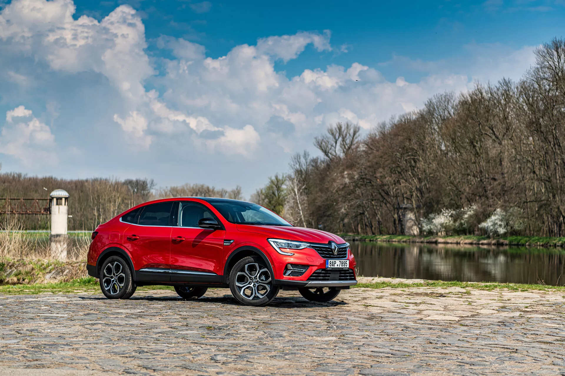 Piros Renault Arkana 2021 SUV parkolva kőburkolaton vízzel és fákkal a háttérben részben felhős ég alatt, oldalnézet kiemeli az elegáns tervezést és dinamikus jellemzőket