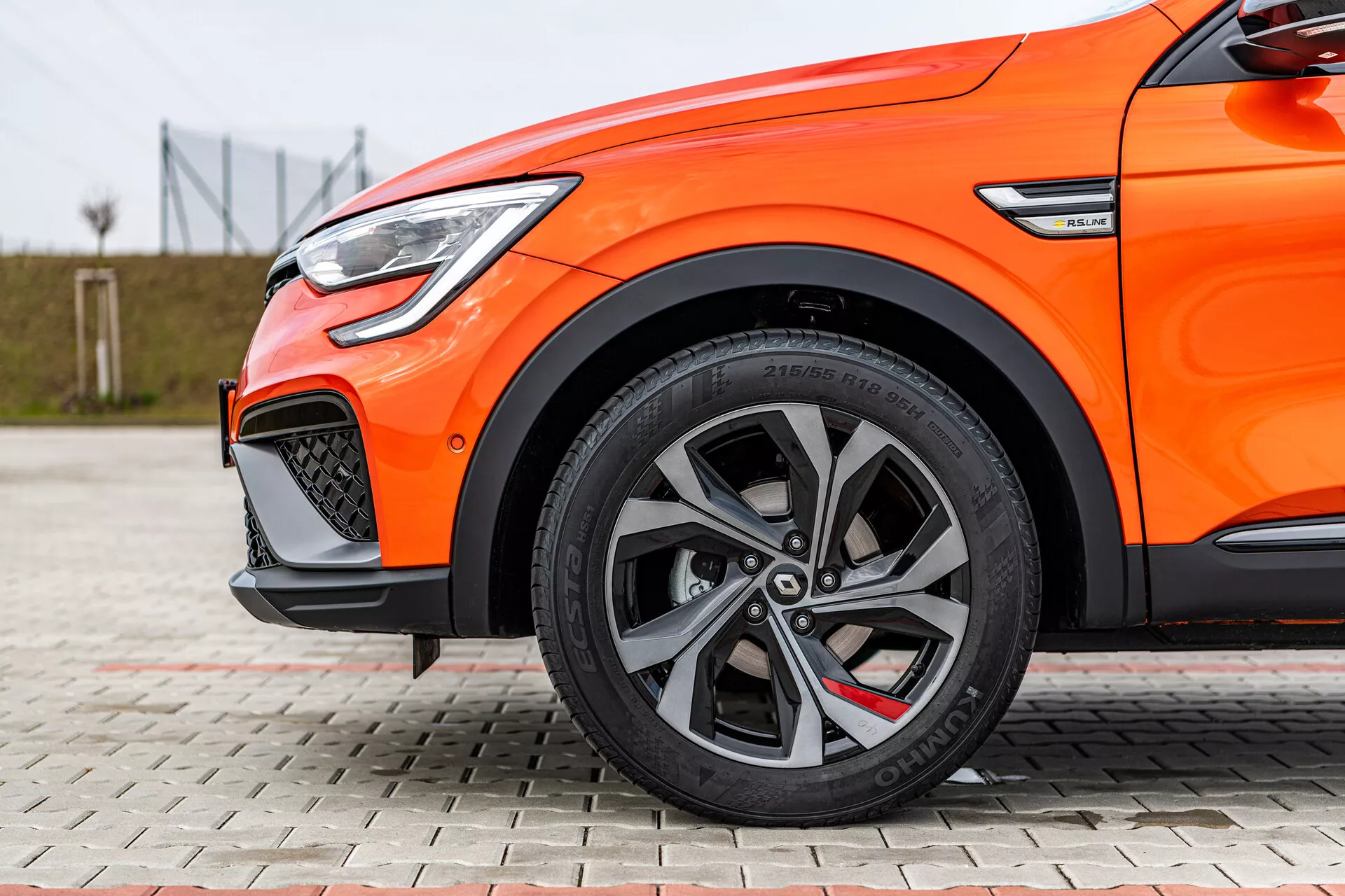 Közeli oldalnézet egy narancssárga 2021-es Renault Arkana autóról, kiemelve a többsugaras ötvözött kerekeket, alacsony profilú gumiabroncsokat, nagy teljesítményű fékrendszert és LED nappali menetfényt