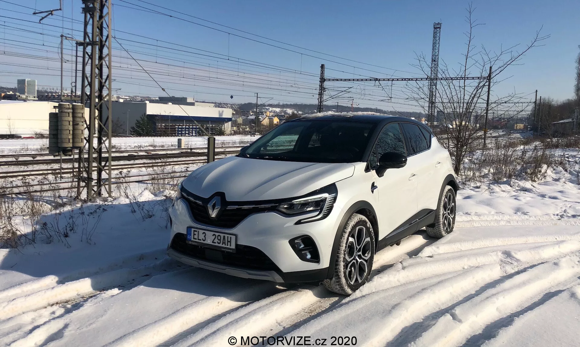 A képen egy fehér Renault Captur 2020, egy kompakt SUV látható, amint egy hóval borított út szélén parkol, háromnegyedes elölnézetből fényképezve. A jármű jellegzetes C-alakú LED nappali menetfényekkel, projektoros fényszórókkal, fekete díszítésekkel ellátott első hűtőrácsával és feltűnő Renault emblémával, valamint többrugós könnyűfém keréktárcsákkal rendelkezik. Figyelemreméltó kialakítási jellemzők közé tartozik a műanyag karosszériaborítás az alsó éleken és a kerékíveken. A környezet hideg, téli napot sugall ipari vagy félig városi háttérrel.