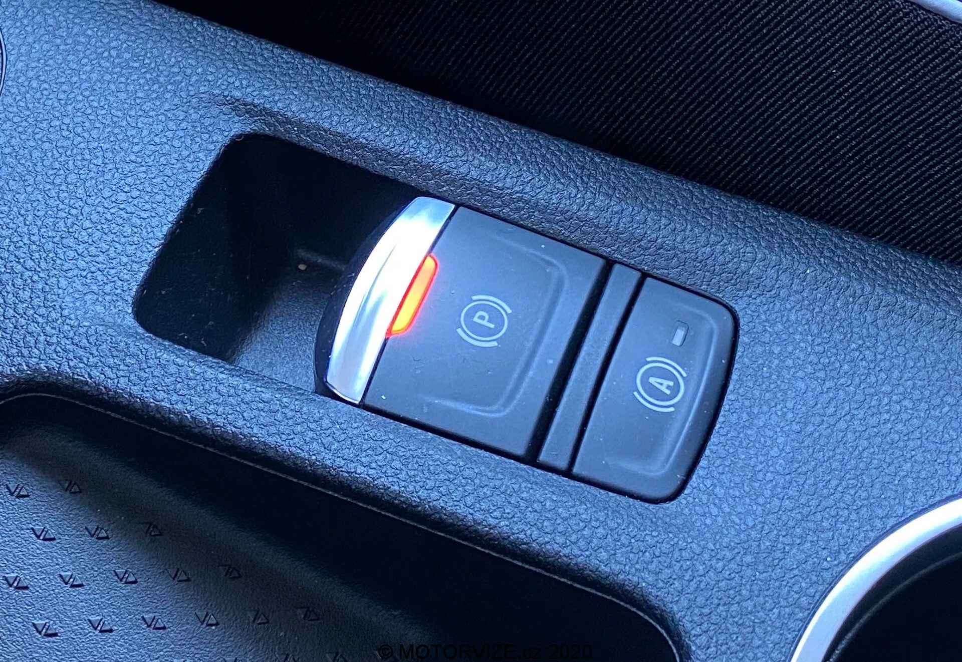 Közeli átlós felvétel a 2020-as Renault Captur belsejében található elektronikus rögzítőfék kapcsolójáról. A középkonzolban elhelyezett kapcsoló két gombból áll: egy pirosan világító gomb zárt lakattal és 'P' szimbólummal, amely az aktív (bekapcsolt) állapotot jelzi, valamint egy másik gomb nyitott lakat szimbólummal. A kapcsoló egy fekete textúrájú műanyag konzolban található, kiemelve a modern járműtechnológiát.