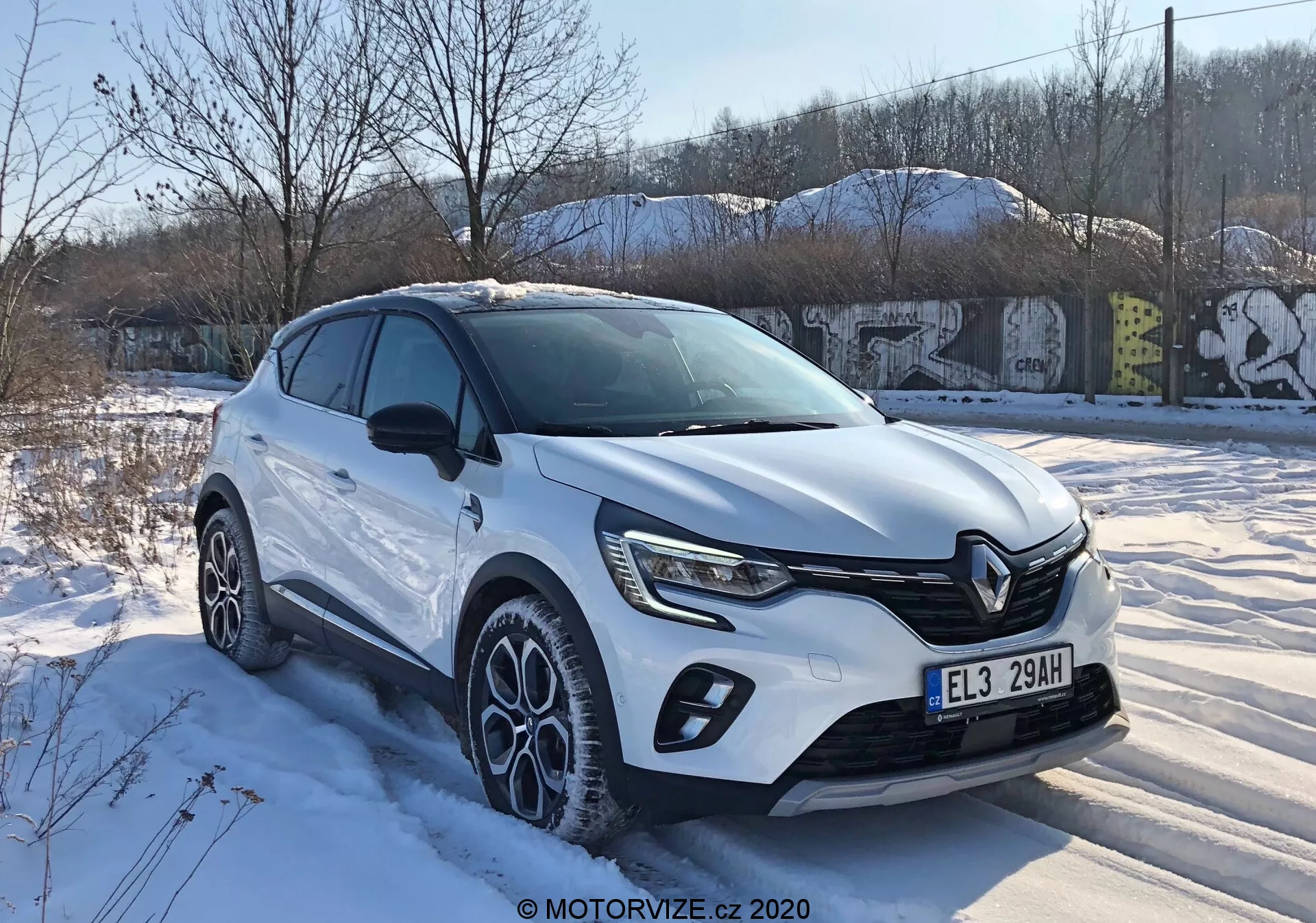 2020-as Renault Captur háromnegyedes elölnézetben, amely bemutatja a modern crossover dizájnt jellegzetes első hűtőráccsal, éles LED fényszórókkal, karosszéria burkolattal, nagyméretű, többküllős könnyűfém kerekekkel és fehér karosszériával havas téli körülmények között városi környezetben.