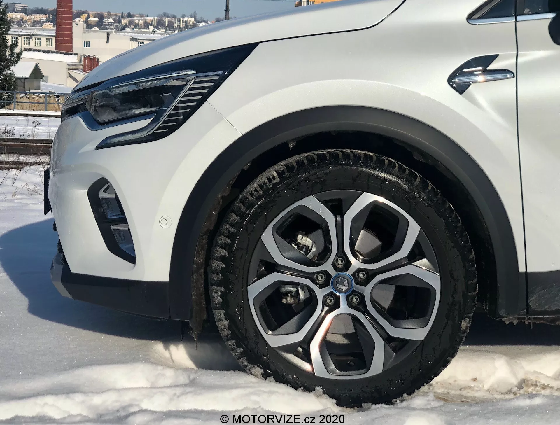 Renault Captur 2020 közeli felvétele alacsony szögből, amely bemutatja a bal első részt, a fókusz az ötvözet keréken, az sárvédőn, a lökhárítón és a fényszóró területén van. Az autó havas felületen áll városi környezetben, épületekkel és tiszta égbolttal.