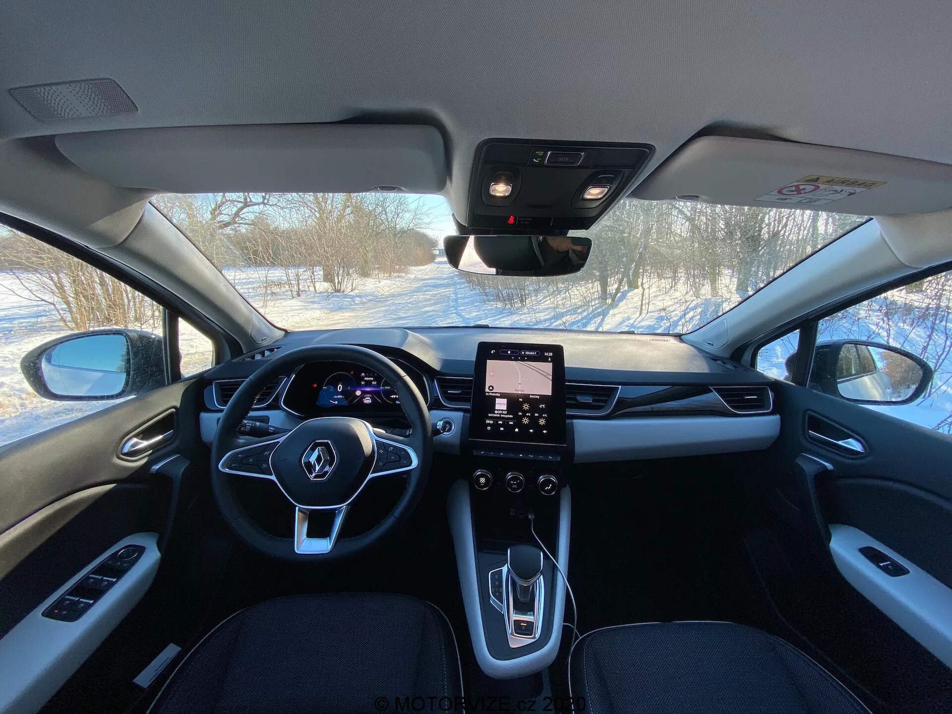 A Renault Captur 2020 belső kép a hátsó ülésekből, amely az elülső kabint mutatja háromküllős kormánykerékkel, integrált gombokkal, digitális műszeregységgel, függőlegesen orientált érintőképernyős infotainment rendszerrel, modern középkonzollal minimális fizikai vezérlőkkel, elegáns váltókar-kerékkel, HVAC szellőzőnyílásokkal, prémium anyagokkal, beleértve a bőrt és fémes díszítéseket, visszapillantó tükörrel, ami valószínűleg ADAS érzékelőket tartalmaz, és az A-oszlop látható, ablakvezérlőkkel.