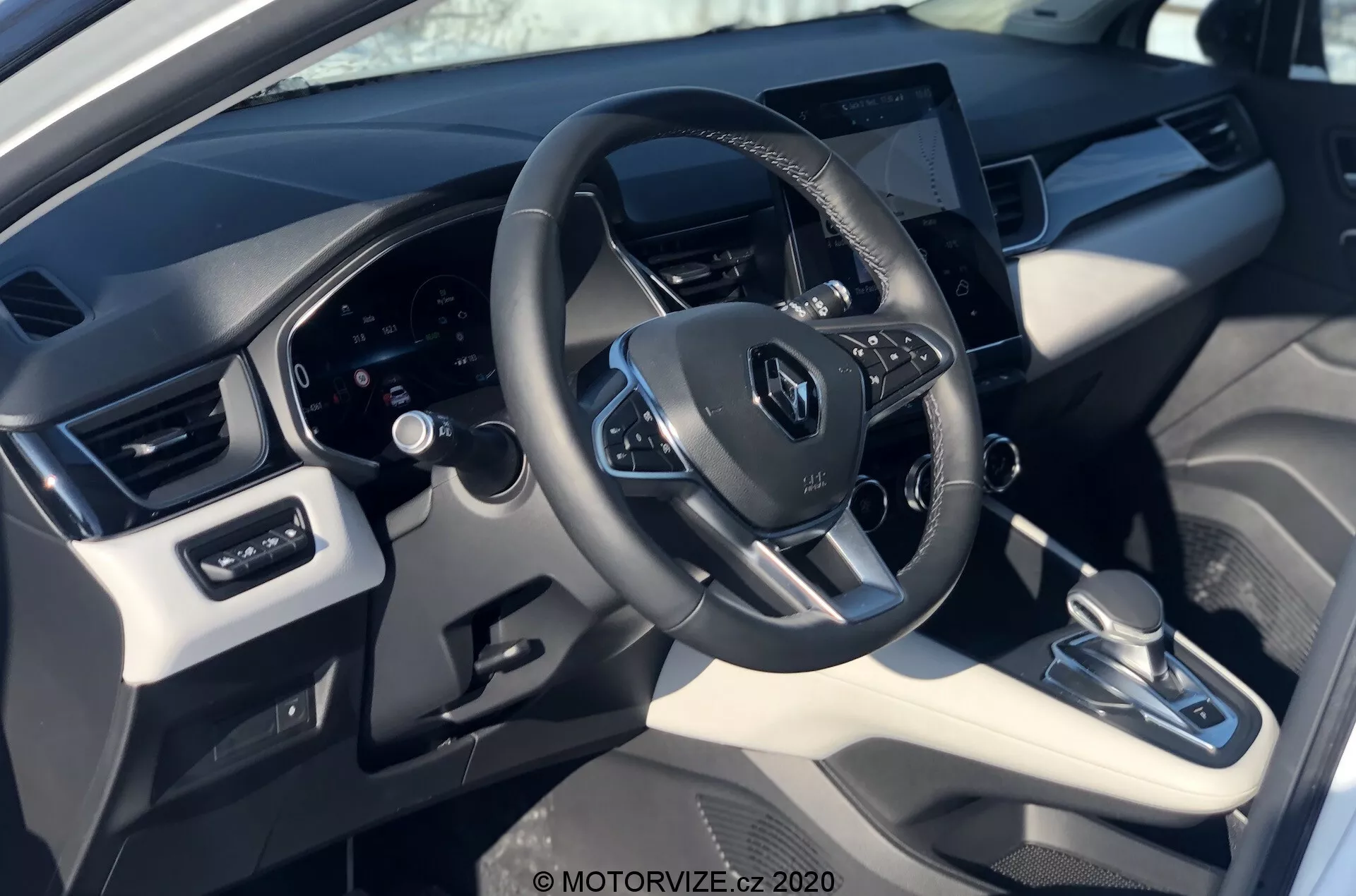 A 2020-as Renault Captur belső nézete a vezető perspektívájából, amely bemutatja a Renault logós kormánykereket, a beépített vezérlőket, a hátul elhelyezkedő digitális műszerfalat és a középre helyezett érintőképernyős infotainment rendszert. A kép kiemeli a szintetikus bőr és műanyag anyagok keverékét fémes ékezetekkel, automata sebességváltóval, klímavezérlő rendszerrel és elektromos ablak- és tükör beállításokkal.