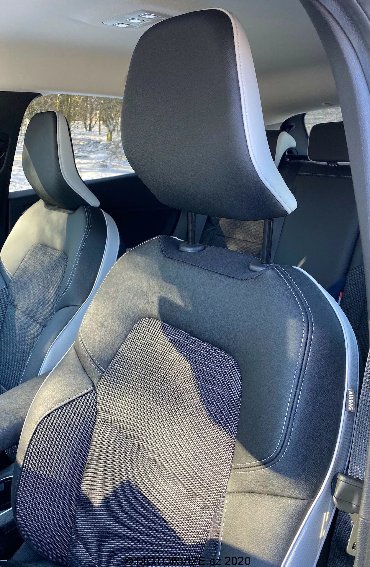 A Renault Captur 2020 belső tere, amely a hátsó részből, kissé jobbról eltolva mutatja az első utasülést és a vezetőülés egy részét. Az ülések felső része integrált fejtámlákkal és látható biztonsági övkeretekkel rendelkezik. Az ülések modern, kéttónusú kialakításúak, sötétszürke és világos szürke színekkel, szintetikus bőr szegéllyel és részletes varrással, amely kiemeli a kézműves munkát és a stílust.