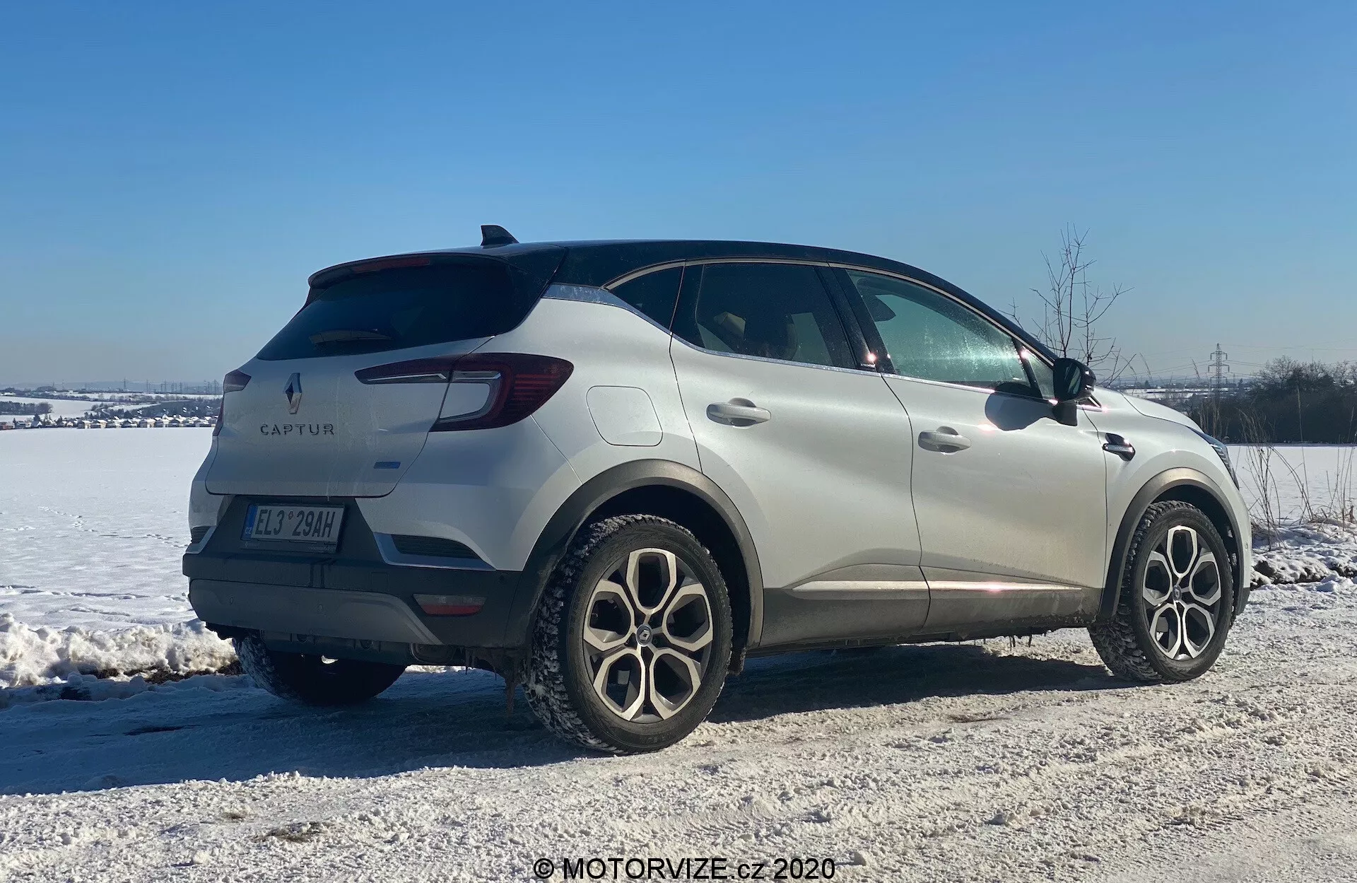 Kép egy 2020-as Renault Captur-ről, egy kompakt SUV-ról, téli környezetben. Háromnegyed hátsó nézetből bemutatva, az autó sima vonalakkal és LED hátsó lámpákkal rendelkező elegáns dizájnt mutat. A gyártó emblémája és a modell neve jól láthatók, mellettük többszárú könnyűfém felnik, hátsó parkolószenzorok és irányjelzővel ellátott oldalsó tükrök. Egy havas, kavicsos területen állva, a kép különféle terepen és téli körülmények között használt jármű képességeire utal.