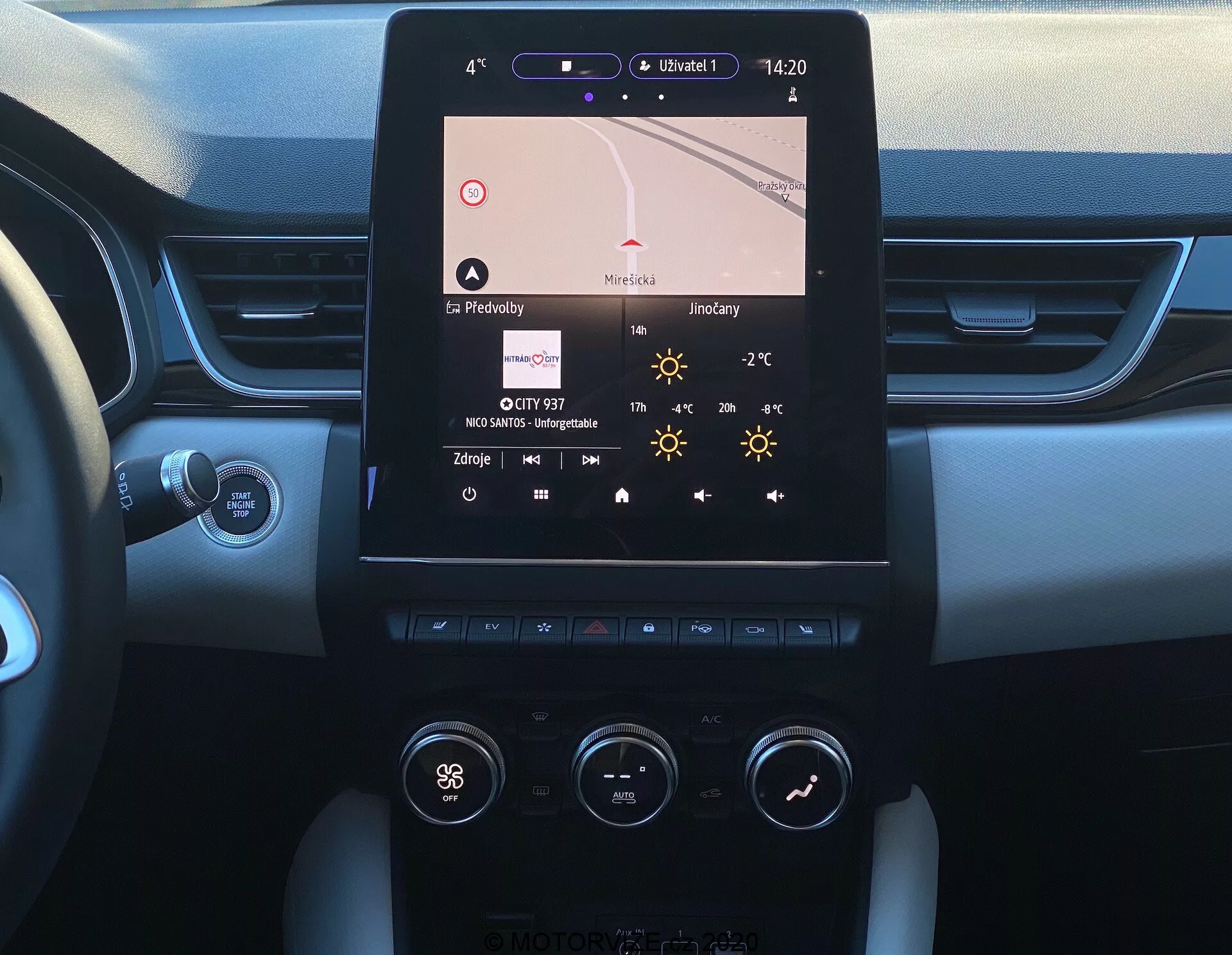 A kép a 2020-as Renault Captur középkonzolját mutatja a vezetőülés perspektívájából. A központi elem egy függőlegesen elhelyezkedő érintőképernyős infotainment kijelző, amely navigációt, időt (14:20), hőmérsékletet (27°C) és rádió részleteket jelenít meg. Alatta látható a kétzónás klímavezérlő rendszer gombokkal és kapcsolókkal, mint például a vezető oldalán 'Hi' hőmérsékleti beállítással. A konzol fém és műanyag anyagok kombinációjával van kialakítva a képernyő és a vezérlők körül. A műszerfal szélei puha érintésű, két tónusú dizájnt tárnak fel, és részben látható a Start/Stop gomb. Látható a kormányoszlop egy vezérlőkarjával, valószínűleg irányjelzésre vagy más funkciókra szolgálva.