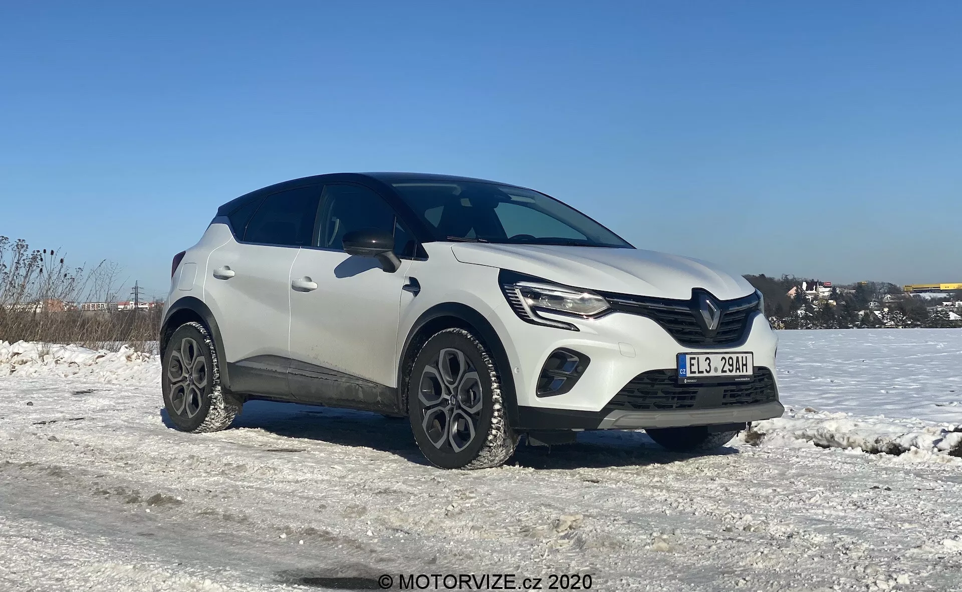 Háromnegyedes nézet egy fehér Renault Captur 2020 kompakt crossover SUV-ról téli környezetben, havas talajjal és tiszta kék égbolttal. A kép kiemeli a jármű bal első oldalát és részleges oldalnézetét, enyhén alacsonyabb szögből készült, kiemelve a kerekeket és az alsó részt. Az autó modern dizájnja jellegzetes vonalakat mutat a motorháztetőn, elegáns LED fényszórókat nappali menetfénnyel és feltűnő első rácsot. Az egyedi könnyűfém kerekek és a kerékjárati ívek körüli műanyag burkolat robusztus megjelenést kölcsönöz. Az autó alatti árnyékok arra utalnak, hogy a fotó délben vagy kora délután természetes nappali fényben készült.