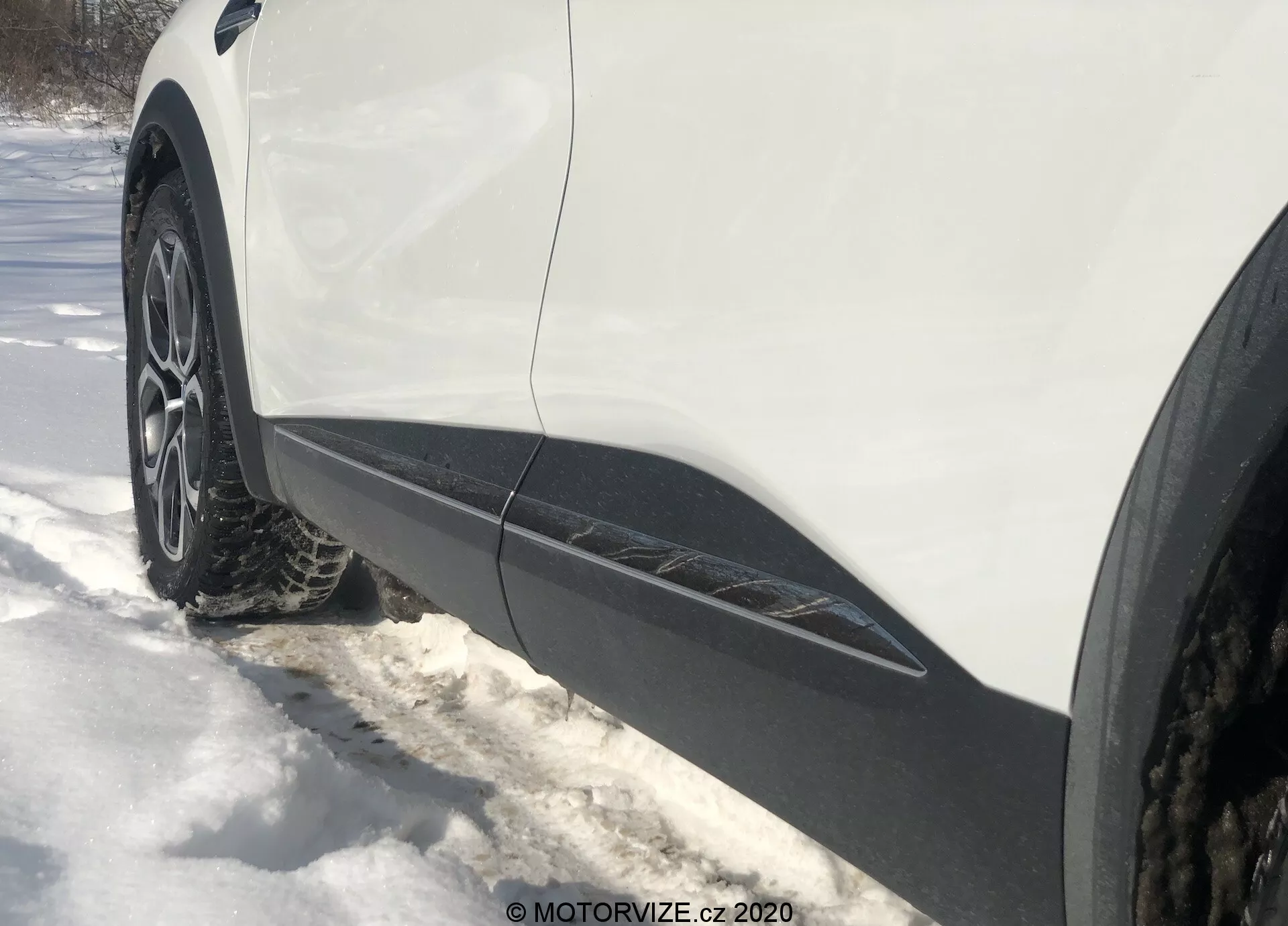 Közelről, oldalnézetből, a föld szintjéről készült fénykép egy 2020-as Renault Captur vezetőoldali első kerekéről és sárvédőjéről. A többküllős alufelni téli vagy négyévszakos abronccsal van körülvéve havas környezetben. A fehér karosszéria fekete díszítéssel szalad az alsó szélen, amit egy crossover vagy SUV jellemzője lehet. A világítás arra utal, hogy a képet napsütéses napon, árnyékokkal készítették.