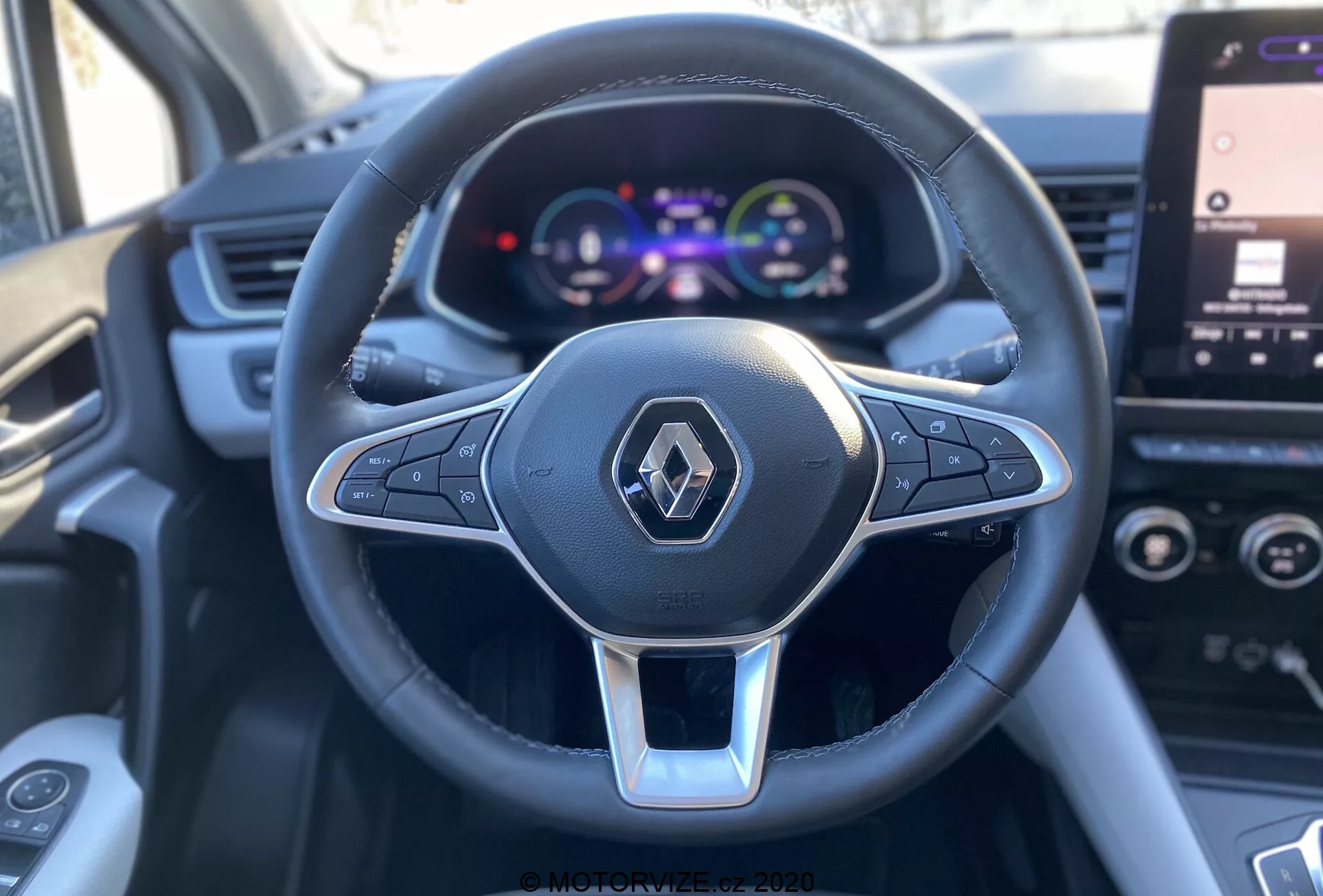 Közelkép, első személy nézete a vezetőülésből egy Renault Captur 2020-es modellben, fókuszálva a multifunkcionális, bőrhuzatú háromküllős kormánykerékre, ezüst díszítéssel és Renault logóval, valamint egy színes digitális műszerfalra, amely a sebességmérőt és a jármű részleteit mutatja, háttérben részben látható infotainment érintőképernyővel.