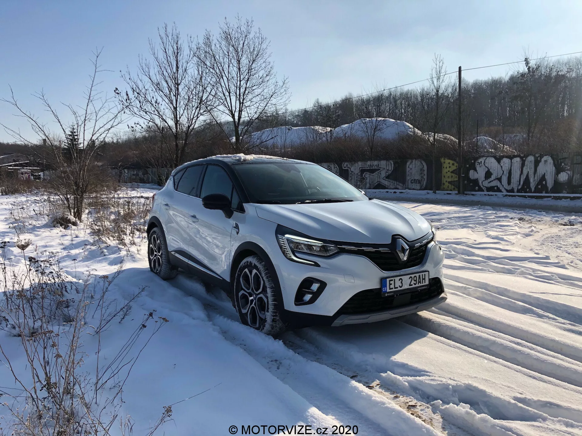 Egy fehér Renault Captur 2020 crossover SUV parkol egy hóval borított talajon, háttérben növényzettel és graffiti borította épületekkel, többnyire derült ég alatt néhány felhővel. A kép egy háromnegyedes elülső szögből készült, a jármű vezetői oldalára koncentrálva, látható a krómos emblémával ellátott hűtőrács és a modern LED-fényszórók. Az SUV elülső LED-ez a megvilágítást, ködlámpa-házakat, karaktervonalakat a motorháztetőn, műanyag borítást a kerékjáratok körül, integrált irányjelzőkkel rendelkező visszapillantó tükröket, többlapátos könnyűfém keréktárcsákat téli gumikkal és erős vállvonalat tartalmaz. A megemelt állás magasabb szabad magasságra utal, amely különböző terepekhez alkalmas.