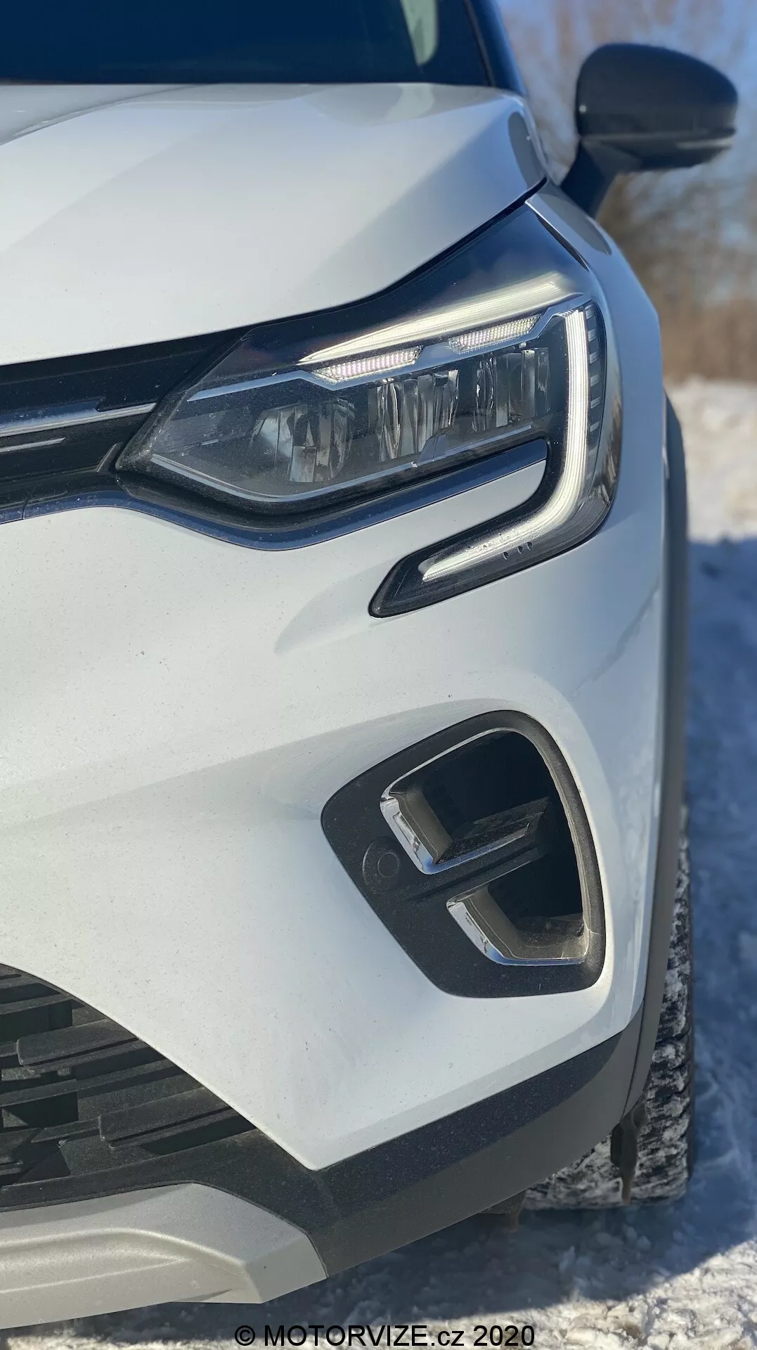 Közelkép egy 2020-as fehér Renault Captur bal elejéről, modern LED fényszóróval, geometrikus elülső lökhárítóval és hűtőrács elemekkel, alacsony elülső szögből készítve, hóval a háttérben