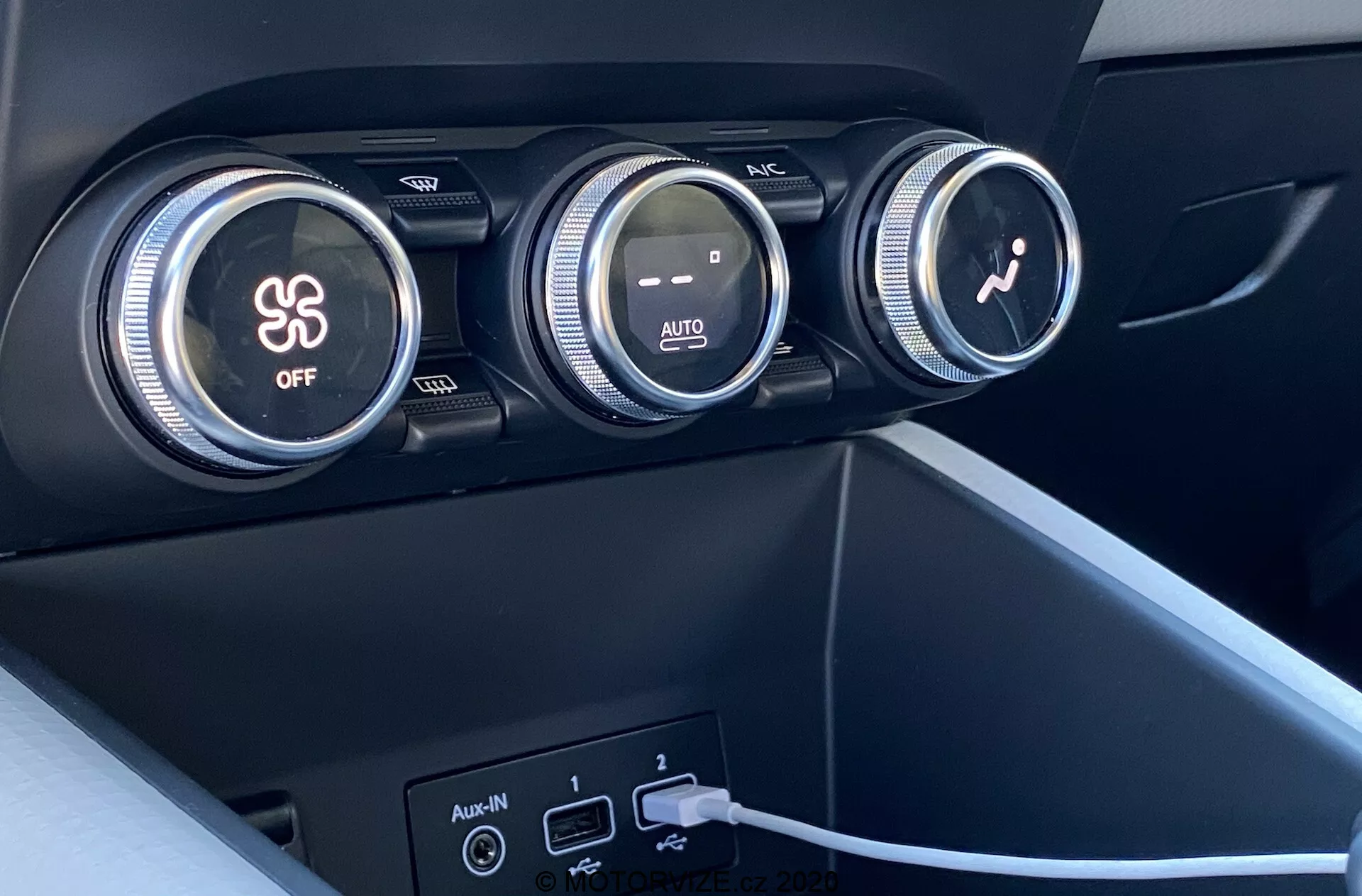 Közelkép a 2020-as Renault Captur belső teréről, különösen a klímaberendezésre koncentrálva. A kép három nagy tárcsát mutat a műszerfalon. Az első tárcsa a légkondicionálót irányítja, hópehely szimbólummal és 'OFF' gombbal jelölve. A középső tárcsa a hőmérsékletet szabályozza, piros és kék színátmenetekkel és 'AUTO' jelzéssel. A harmadik tárcsa egy ventilátor ikonnal a ventilátor sebességét állítja be. A tárcsák alatt két USB-port és egy segéd bemenet található, az egyik USB-porthoz csatlakoztatott kábellel. Az anyagok matt felületű sötét műanyagból vagy szintetikus anyagból készültek, és modern, minimalista dizájnnal rendelkeznek.