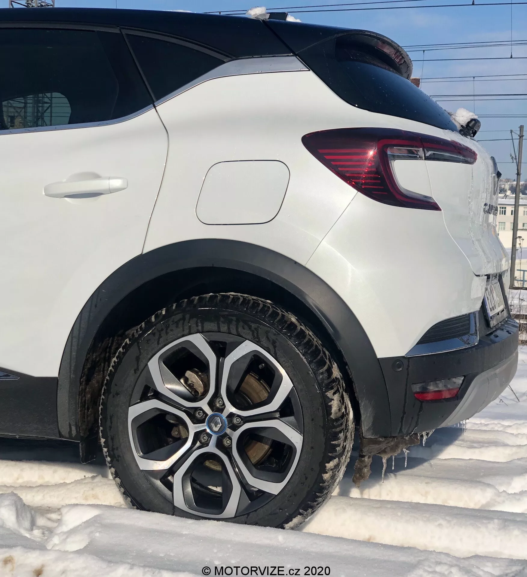 Részletes közeli kép egy fehér 2020-as Renault Capturról, amely az utasoldal hátsó részét mutatja alacsony szögből. Kiemeli a sokküllős alufelnis hátsó kereket, amelyet sár és hó takar, jelezve téli körülményeket. A kép tisztán rögzíti a hátsó kerékívet, a modern hátsó lámpát vörös és átlátszó LED szegmensekkel, és a kerék mögötti robusztus műanyag burkolatot. A hátsó csomagtérajtó ferde ablaka hozzájárul a jármű aerodinamikai formájához.