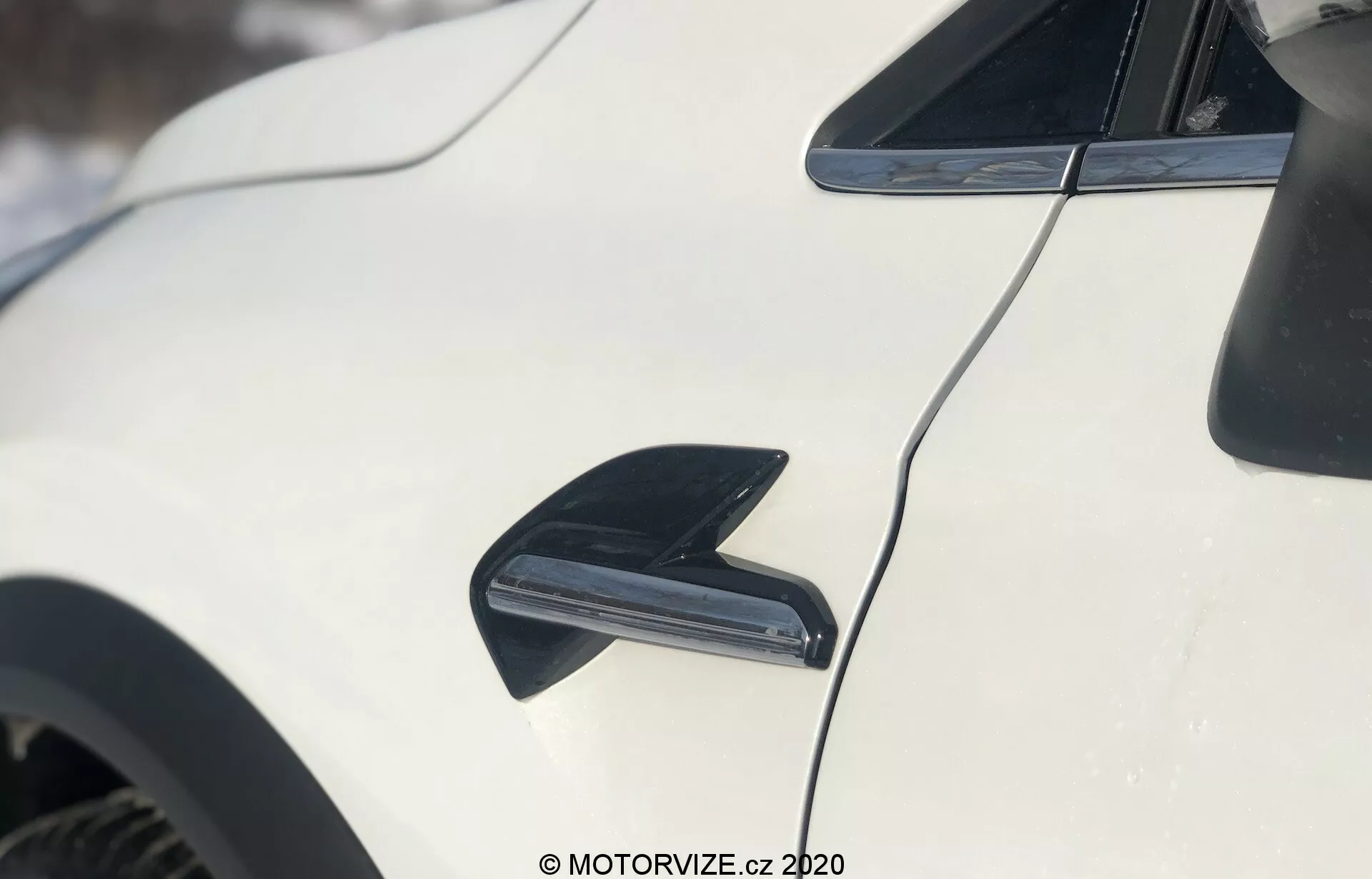 Közelkép a Renault Captur 2020-as modelljének fehér külsejéről, a jármű oldalára összpontosítva. A kép bal felső sarkában egy fekete, cápauszony formájú antenna látható, amely a rádiójel vételére és az aerodinamikára szolgál. Az antenna alatt középen egy fekete szellőzőnyílás vagy légbeömlő csatorna emelkedik ki, ami valószínűleg a fékek hűtésére vagy a légáramlás javítására szolgál. A jármű karcsú, aerodinamikus tetővonala hátrafelé ível, a fényes fehér festés pedig visszatükrözi a fényt. A háttér enyhe elmosódása hóborította külső környezetre utal.