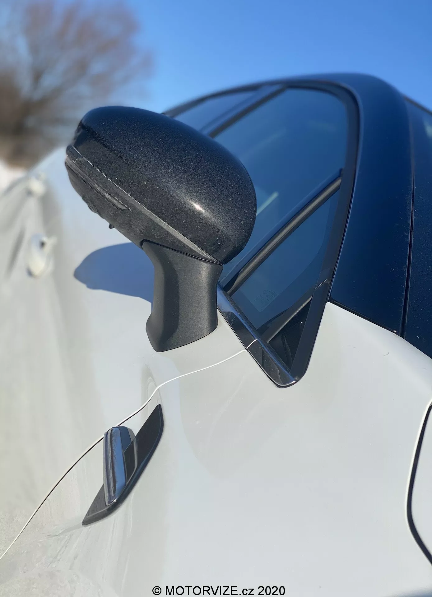 Az Renault Captur 2020 oldalsó tükör szerelvényének közelképe elölről hátra, a fekete texturált műanyag tükörházra összpontosítva, beépített irányjelzővel és a kék ég tükrözésével. Az alatta lévő elegáns sötét színű karosszéria panel és a lehetséges holttér tükör modern dizájnt és fejlett funkciókat jelez.